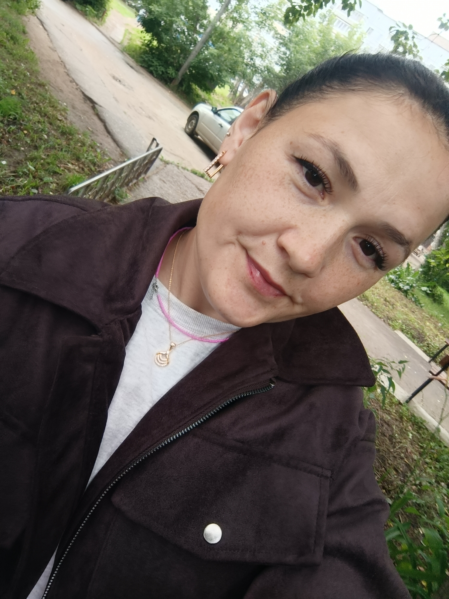The image shows a woman posing for a selfie outdoors. She has fair skin with freckles and dark hair pulled back. She is wearing a dark brown jacket, a light-colored shirt, and a necklace with a crescent moon pendant. The background shows a paved road, some greenery, and a silver car parked on the side of the road. The presence of a silver car and the overall environment suggests an urban or suburban setting. Given the mention of Chaikovsky, Russia, it is likely this scene is set in that city, with the location being a street or roadside area.