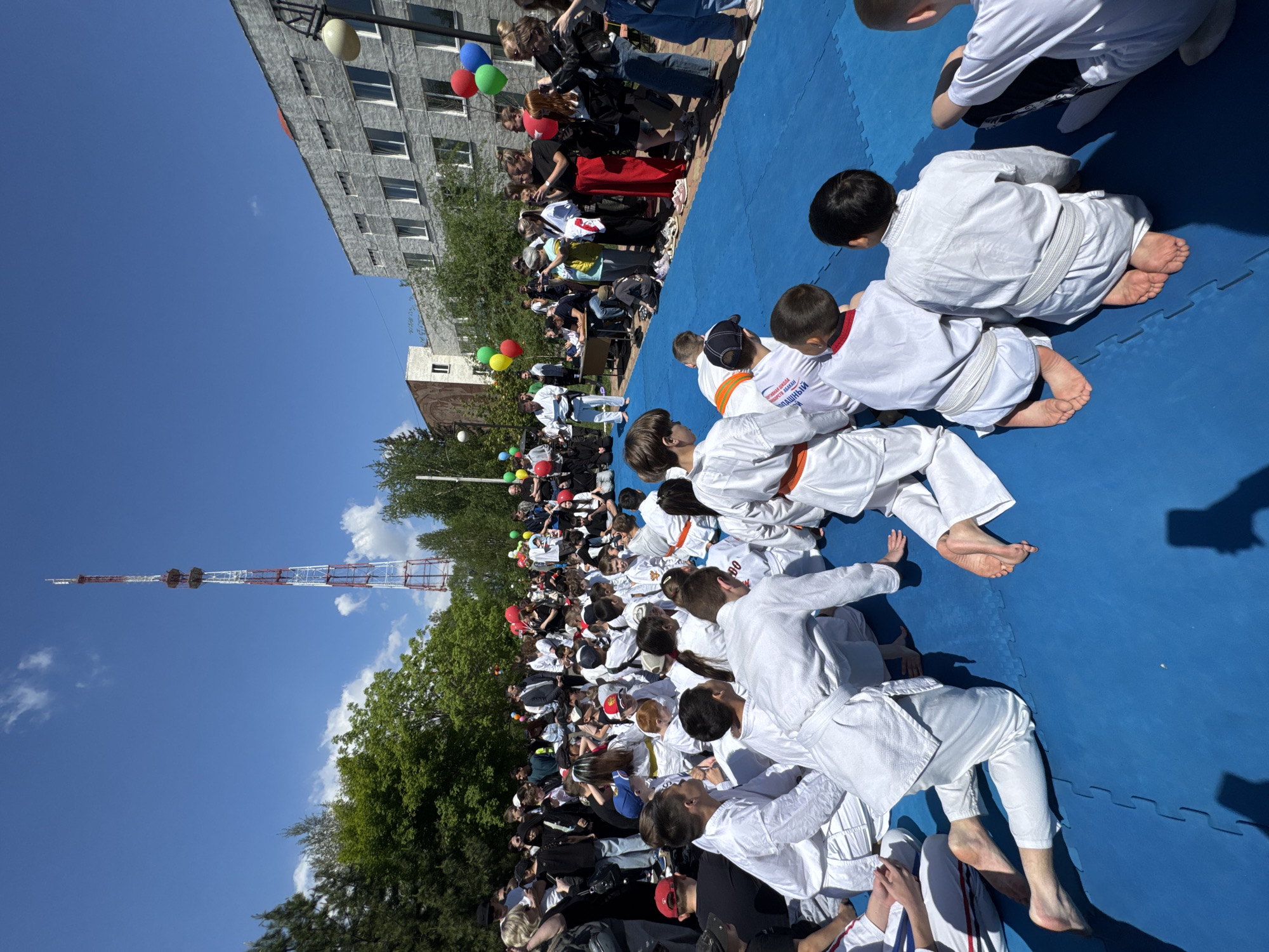 The image shows a group of people, mainly children, dressed in white martial arts uniforms on blue mats outdoors, likely participating in a martial arts event or class. They are in Abakan, Russia, near the Abakan TV Tower, based on the tower visible in the background. A crowd of onlookers, including adults and children, is gathered around the mats. The environment appears to be a public space. The image was taken on a sunny day.