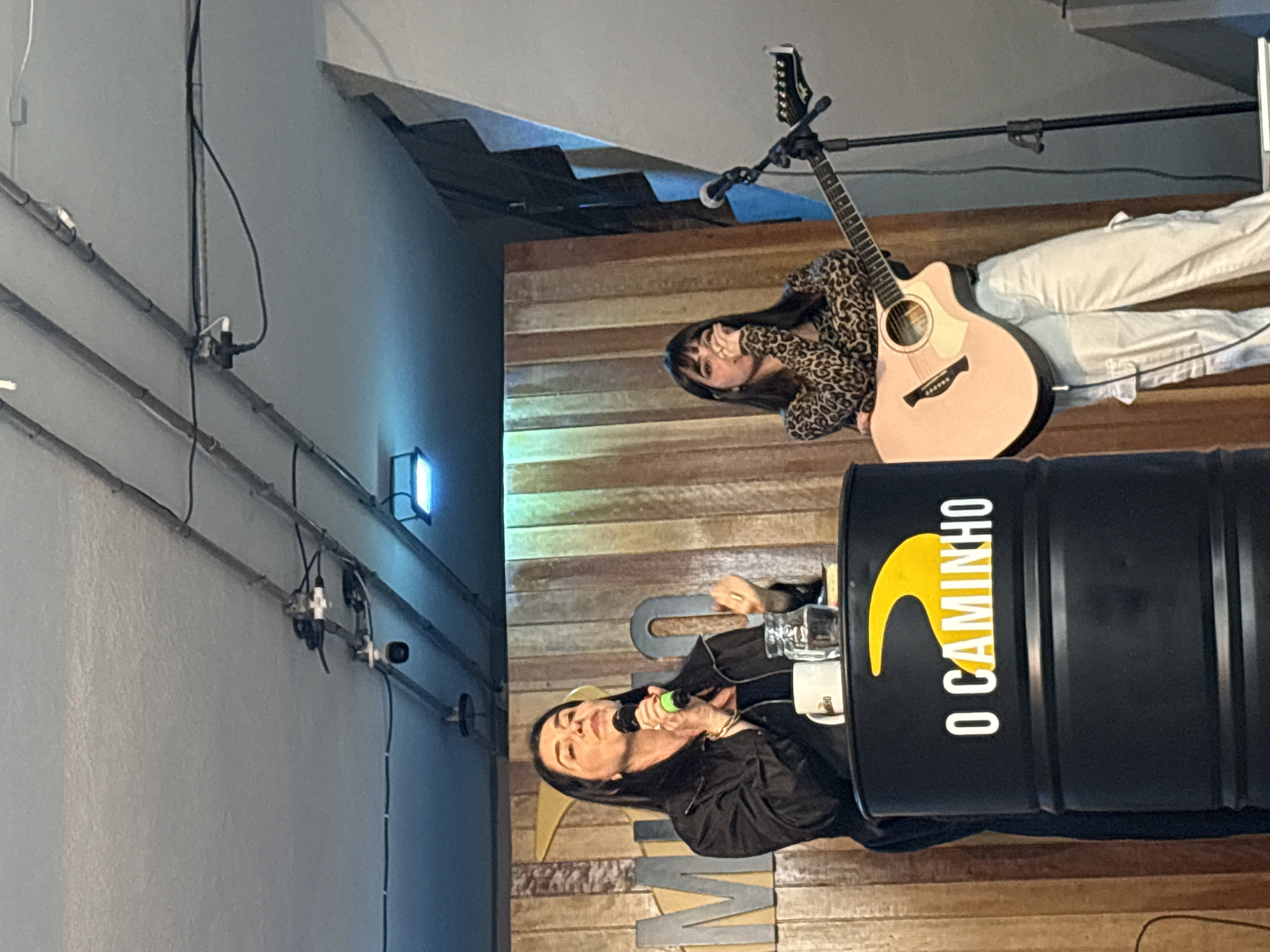 The image depicts an indoor event, possibly a church service or a similar gathering, with two women on a stage. The woman on the left is speaking into a microphone, holding it with her right hand, and gesturing with her left hand. She is wearing a black jacket over a black top and has dark hair. She is standing behind a black cylindrical object with yellow text and a yellow symbol that reads "O CAMINHO." A white mug and a glass of water are placed on top of this object.
The woman on the right is holding a pink acoustic guitar and appears to be singing or preparing to sing. She is wearing a leopard-print top and white cargo pants. A microphone is positioned in front of her. The background consists of wooden planks and a gray ceiling with visible electrical wiring and some lighting fixtures. The overall setting suggests a casual and intimate performance or presentation space. The specific location in the city of São Paulo, Brazil cannot be determined from the visual information provided.