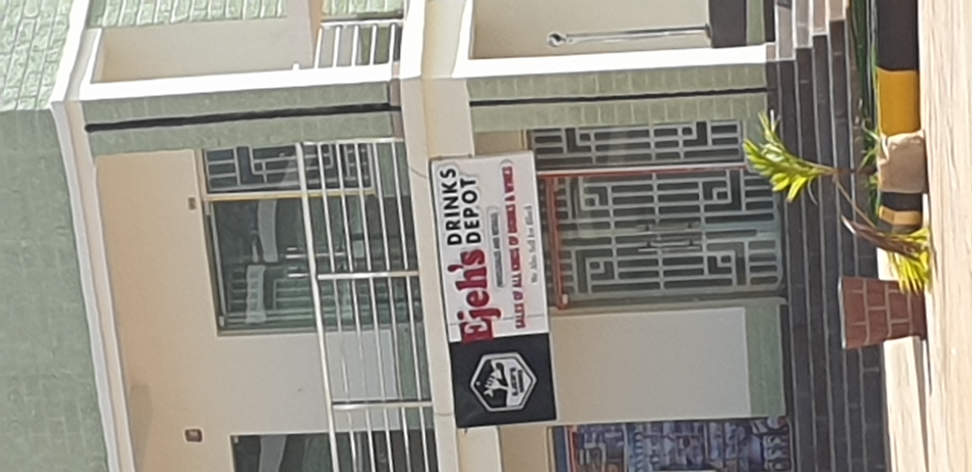 The image shows the exterior of a building identified as Ejehs Drinks Depot, located in Sabon Tasha, Nigeria. The building has a light-colored facade with green brick accents on the upper portions and around the corners. A balcony with a silver railing is visible on the second floor.
A prominent sign for "Ejehs DRINKS DEPOT" is displayed on the front of the building, featuring a logo with a hexagonal shape and stylized imagery. Below the main text, smaller text reads "SALES OF ALL KINDS OF DRINKS & WINES". Another line of text states "We Also Sell for Black".
The ground floor features a set of double doors with intricate metal grates in a geometric pattern. To the left of the doors, there is a glimpse of a display inside, partially obscured by a white wall.
In the foreground, there are several decorative elements. A red brick planter holds a small palm plant with some fallen fronds. In front of the plant, there are two planters, one with a burlap-like covering and another with yellow and black banding. Steps lead up to the entrance of the building. The overall scene suggests a commercial establishment in an urban setting.