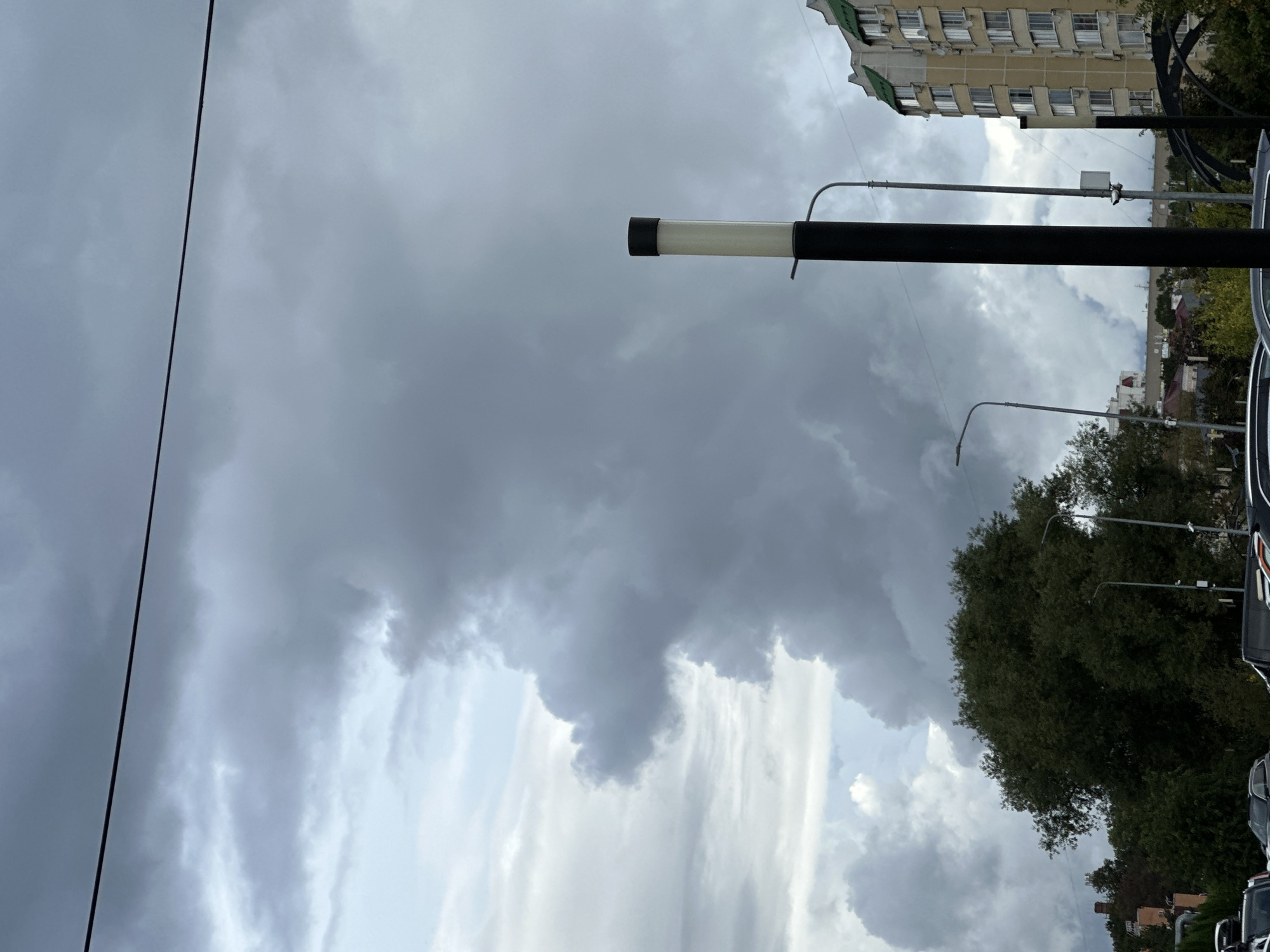 The image depicts a cloudy sky over a city, possibly Podolsk, Russia. A prominent, dark gray cloud formation dominates the upper portion of the frame, with lighter clouds visible in the background. In the foreground, a black lamppost with a white illuminated cylinder stands to the right of the frame. To the right of the lamppost, a multi-story building with light-colored walls and green accents on the roof is partially visible. Below the lamppost and building, a dense green tree is seen. The lower part of the image shows the tops of several parked cars and what appears to be a street or parking lot. A thin wire stretches across the top of the image. The overall mood is overcast and somber, suggesting inclement weather.