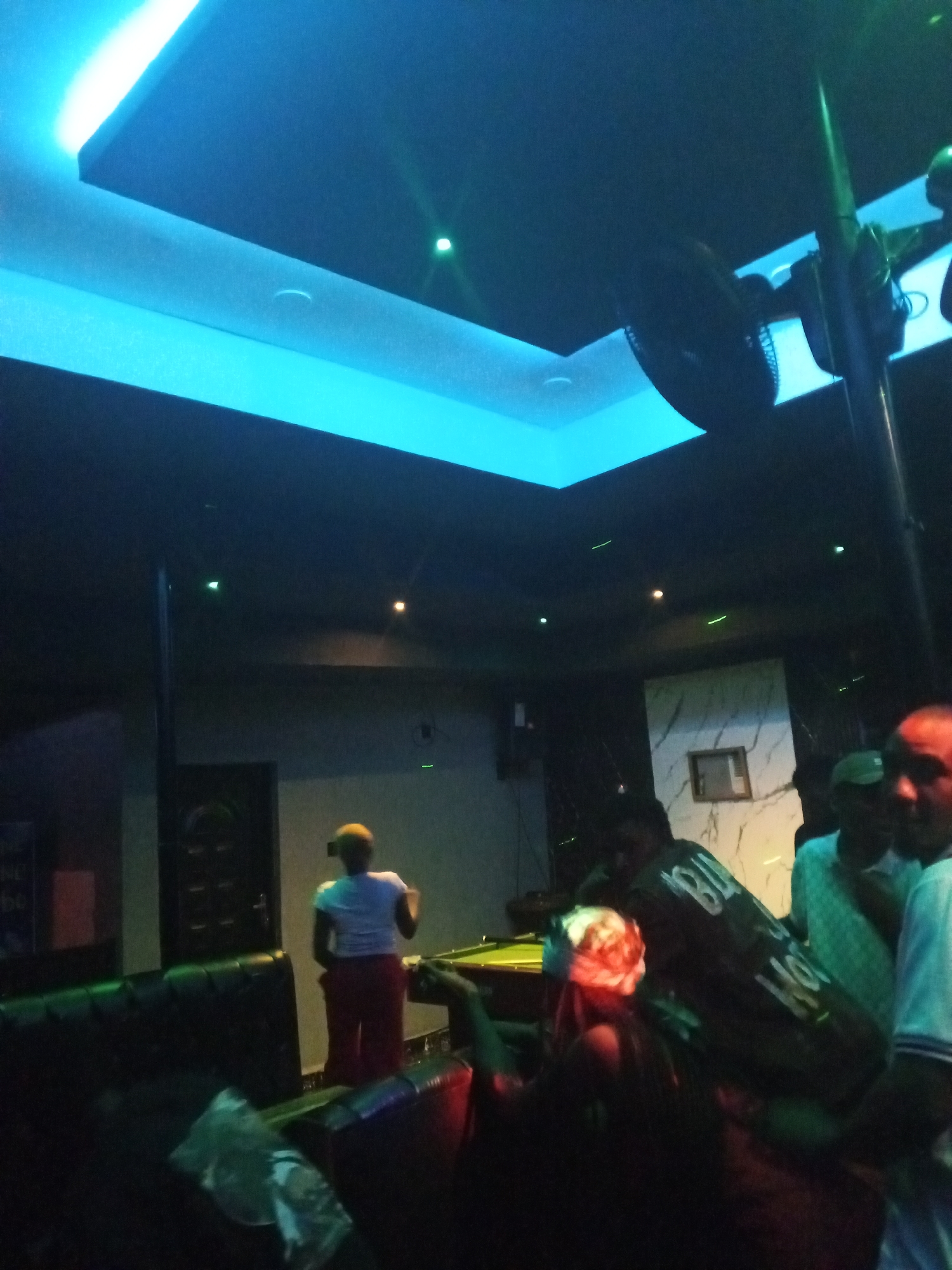 This image depicts an interior scene, likely a bar or lounge, characterized by its dim, atmospheric lighting. The setting appears to be an entertainment venue in Lokoja, Nigeria, at night, given the reliance on artificial light.
The ceiling features prominent cool blue LED strip lighting in recessed panels, along with small green and orange/yellow spot lights scattered across the dark surface. Large, dark ceiling fans are visible, including one on the right mounted on a pole, suggesting a need for air circulation.
Several people are present. In the mid-ground, a person wearing a white shirt and red/maroon pants, with a yellow head covering, stands with their back to the camera, near a green-felt pool table. This person is positioned in front of a dark door and a wall paneled with marbled material, featuring a framed item. In the foreground, dark, upholstered sofas are visible.
To the right, a group of people is partially captured; one individual on the far right has a reddish hue on their face, and another beside them wears a green cap. Another person in the mid-ground wears a dark shirt with white text, where "BLOOD" is partially legible. The activity appears to be social interaction, possibly including games like pool, within a relaxed, dark-lit environment. On the far left, a partial sign displays "NE" and "60".
