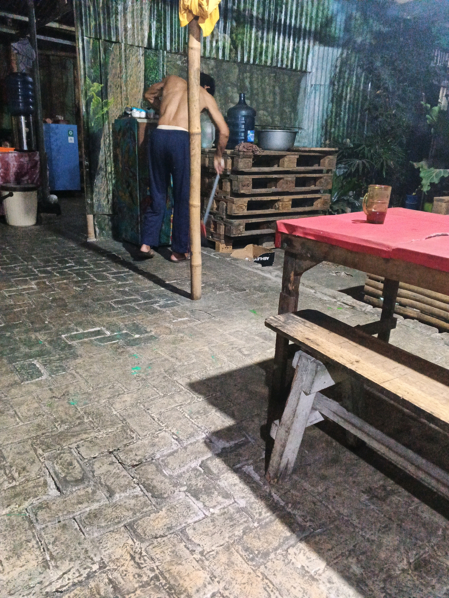 The image shows a person sweeping a tiled outdoor area. The person, facing away from the camera, is wearing dark pants and is shirtless. They are holding a broom and appear to be cleaning a floor made of interlocking rectangular tiles. The scene is dimly lit, suggesting it is either dusk or nighttime.
In the background, there are several objects and structures. To the left, there is a large blue refrigerator or cooler and a water dispenser with multiple blue bottles. Behind the person sweeping, there is a corrugated metal wall with a printed pattern resembling trees and foliage. A stack of wooden pallets is visible, topped with a metal basin and a large blue water bottle. Further to the right, a red-covered table with a wooden bench in front of it is present. A glass with a reddish liquid sits on the table.
The environment appears to be an outdoor area, possibly a backyard or a service area, characterized by the tiled floor, the presence of utilitarian items like the water dispenser and pallets, and the corrugated metal wall which might be part of a shed or an attached structure. The overall impression is one of a functional, possibly semi-industrial or residential outdoor space. The mention of Ungaran, Indonesia, suggests this is the geographical context for the scene.