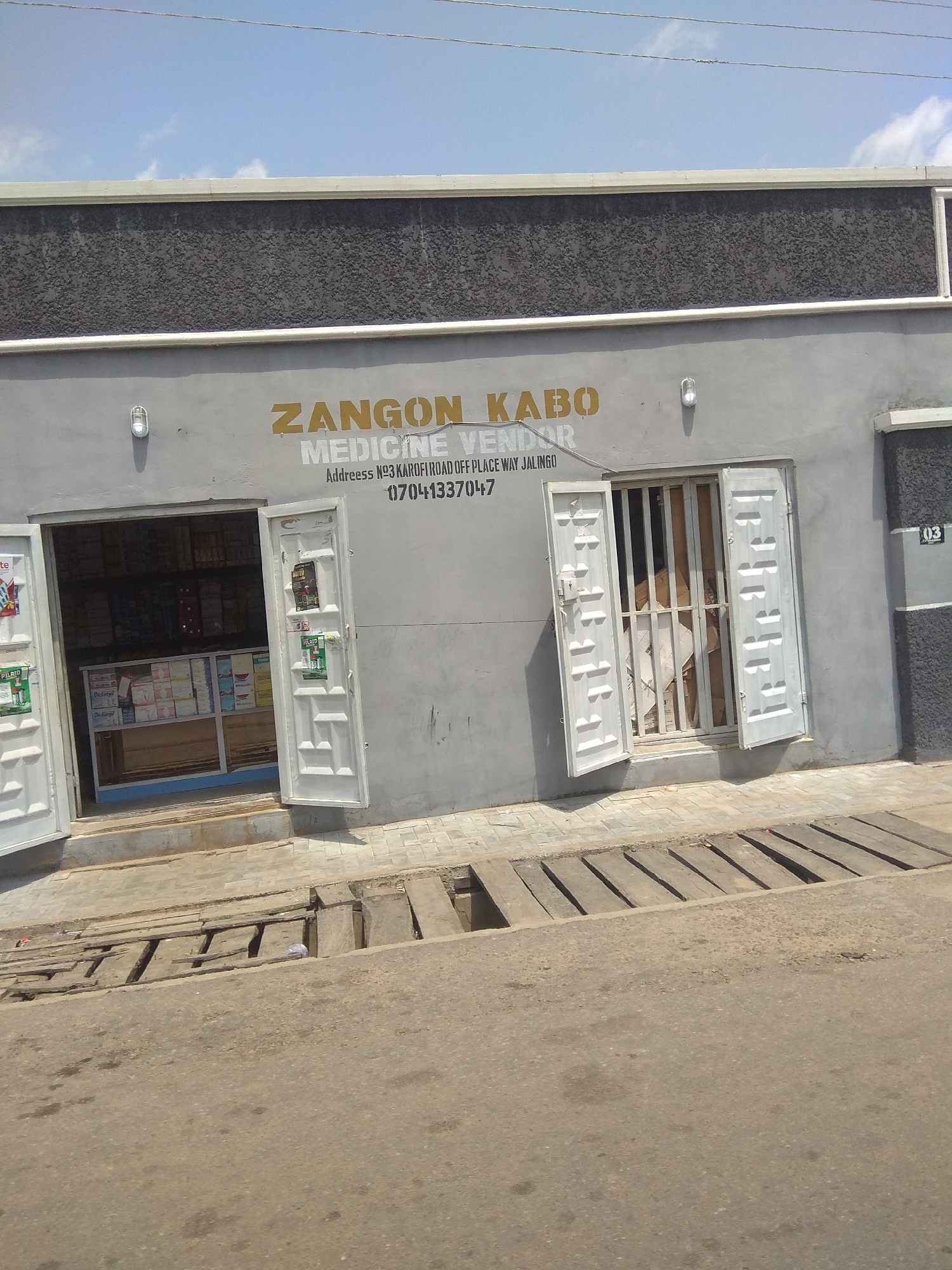 The image shows the exterior of a business named "ZANGON KABO MEDICINE VENDOR" located at Address No3 KAROFI ROAD OFF PLACE WAY JALINGO. The building is light gray with a dark textured band across the top. Two white double doors are open on the left side, revealing shelves stocked with boxes of medication. Another set of white double doors on the right are also open, covering a barred window. The street in front of the building is paved with concrete tiles, and a section of the road has a wooden cover over a drainage ditch. The sky is blue with a few white clouds, and power lines are visible across the top of the frame.