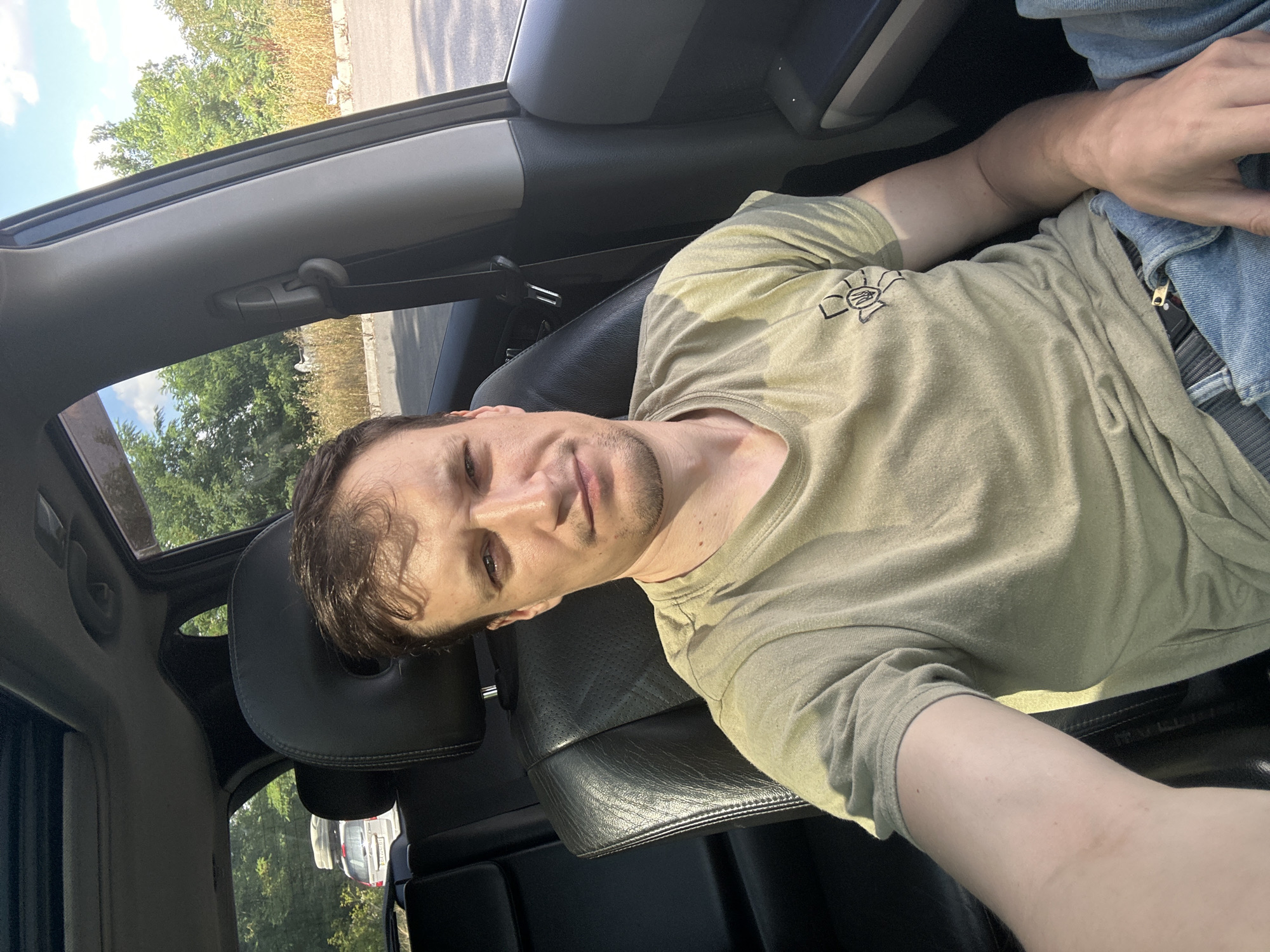 A man with short brown hair and light skin is sitting in the driver's seat of a car, taking a selfie. He is wearing a light green t-shirt with a black emblem on the left breast. He is also wearing blue jeans. The car has black leather seats and a sunroof. Outside the car, there are trees and dry grass. A silver SUV with a roof rack is visible in the background. The location is Dunaivtsi, Ukraine.