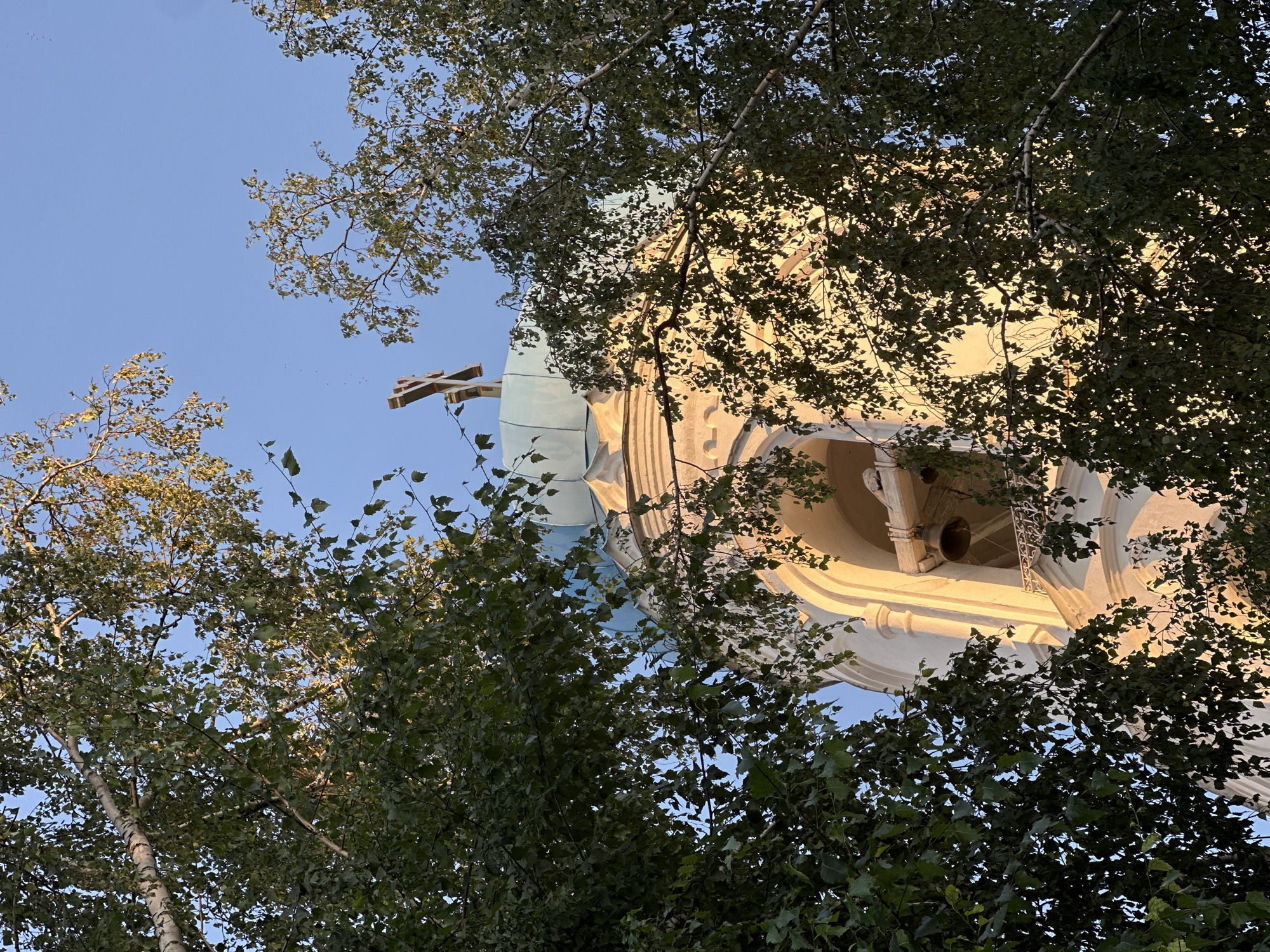 The image shows the upper part of a church bell tower, partially obscured by the branches and leaves of trees. A cross is visible atop the dome of the tower, which is light yellow and capped with a pale blue dome. The bell tower itself is white with arched openings. A bell is visible hanging within one of these arches. The sky is clear and blue. This appears to be a view of the bell tower of the Assumption Cathedral in Biysk, Russia, judging by the architectural style and the presence of a distinct cross and dome.
