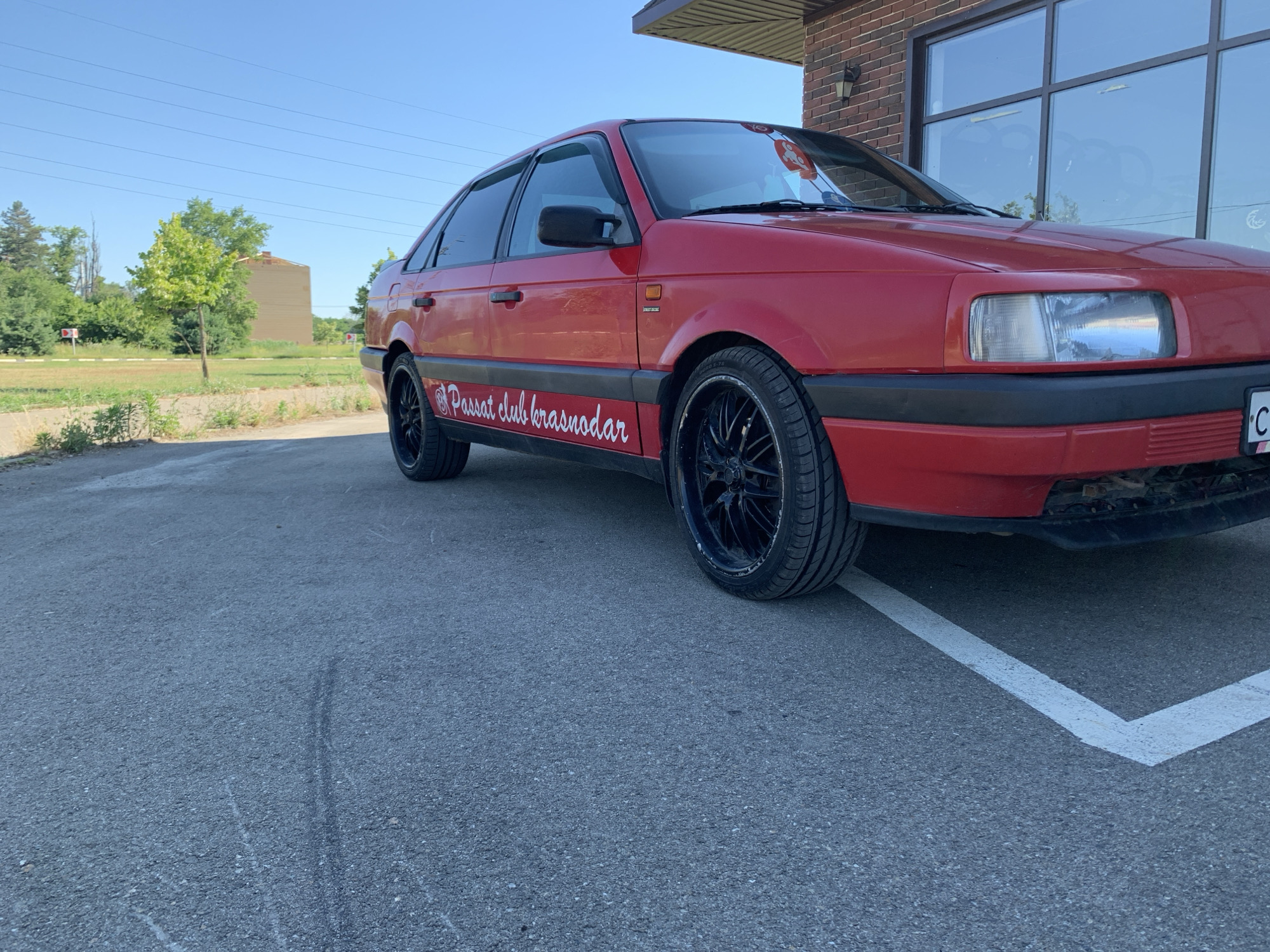 A bright red sedan, a Volkswagen Passat, is parked on an asphalt surface, possibly a parking lot, in Kropotkin, Russia. The car has black rims and a decal on its side that reads "Passat club krasnodar". The car is positioned at an angle, with the front passenger side wheel closer to the viewer. The background features a brick building with large windows, a clear blue sky, and some greenery including trees and a building in the distance. The lighting suggests it is daytime, with sunlight casting shadows. There are white lines painted on the asphalt, indicating parking spaces. The overall scene is static, with no people visible.