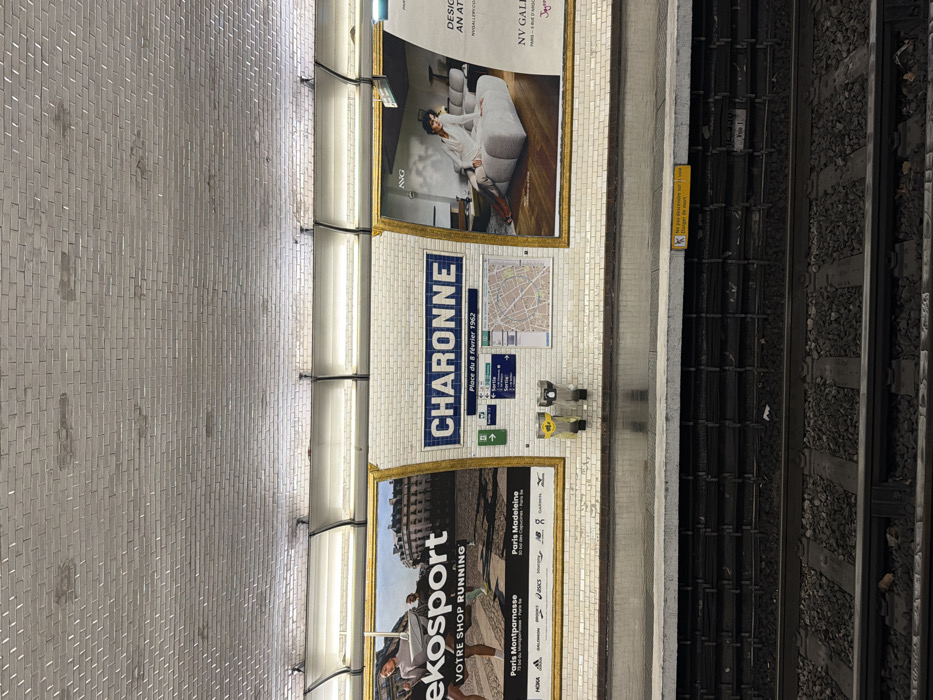 The image captures a metro station platform at Charonne in Paris, France. The scene is illuminated by multiple overhead fluorescent lights positioned above the tiled wall. The station name "CHARONNE" is centrally displayed in blue lettering on white rectangular tiles, framed by a blue border. Below the station name, a plaque reads "Place du 8 Février 1962," flanked by a green and white exit information panel on the left and a detailed city map of Paris.
Two large advertisements, each enclosed in a gold frame, dominate the wall. The left advertisement for "ekosport" features people running and lists store locations in "Paris Montparnasse" and "Paris Madeleine," along with various sports brand logos. The right advertisement for "NV GALLERY" showcases a woman sitting on a modern white sofa. Beneath the central station signage, two wall-mounted, light-yellow dispensers are visible.
The platform is made of grey concrete, showing a reflective surface. A yellow warning sign on the platform's edge, visible on the right, states "Ne pas descendre sur la voie" and "Danger de mort." Below the platform, two metro tracks are laid on a bed of dark gravel ballast, separated from the platform by a tiered, dark-colored structure. The arched ceiling of the station is covered in small white rectangular tiles, some of which exhibit discoloration and minor surface wear. No individuals are present on the platform.