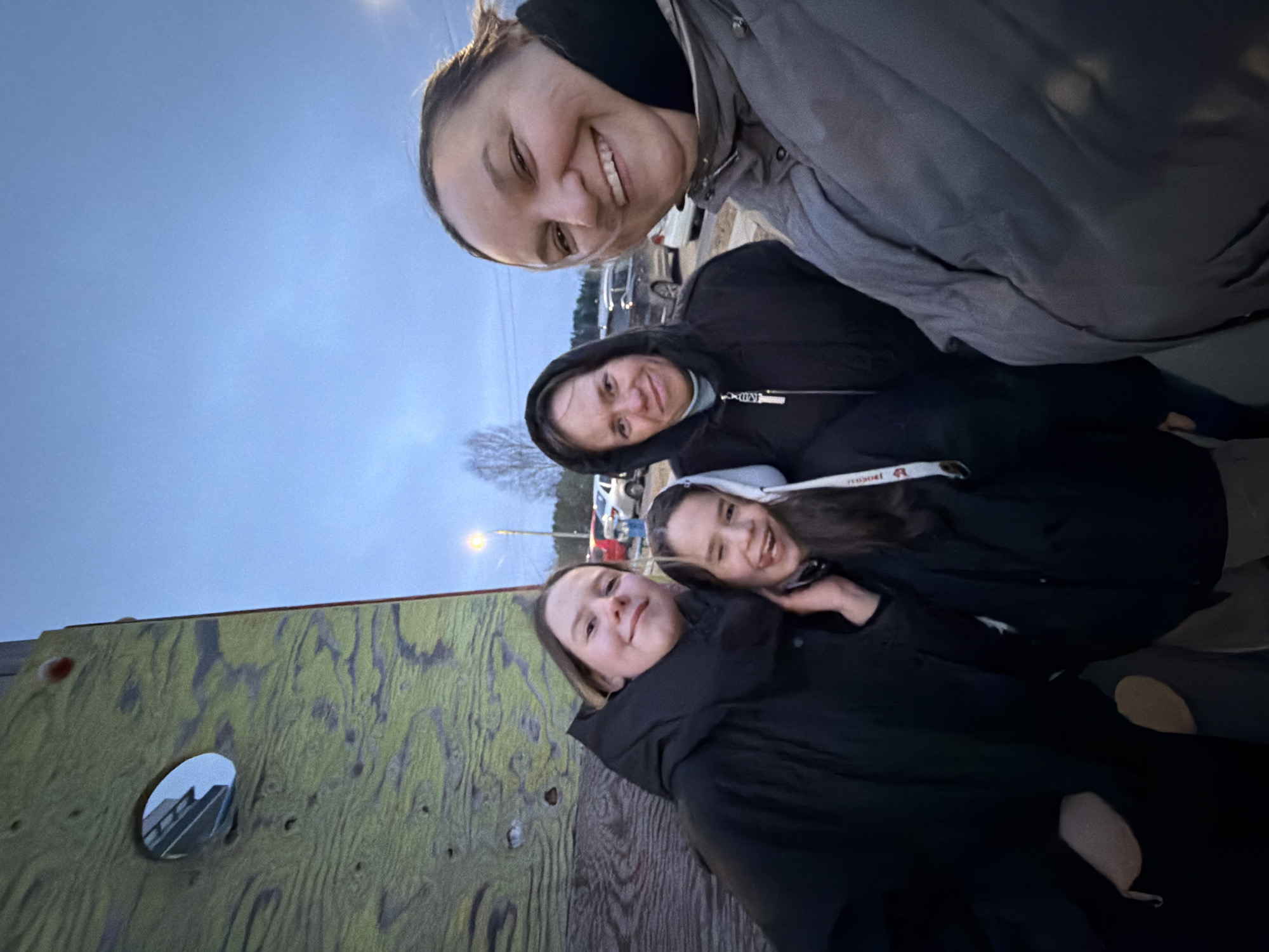 This outdoor group photo, taken in Chaikovsky, Russia, features four females, likely family or friends, posing closely together with smiles. The image is oriented vertically, with the camera held at a low angle looking upwards.
The person closest to the camera, positioned at the top of the frame, is an adult woman with dark hair and a wide smile, wearing a grey jacket. Below her, another adult woman in a black hooded jacket smiles towards the camera. Further down the frame are two younger females. One young woman with dark hair, wearing a black hooded jacket with a visible white drawstring, is smiling. Her arm is around the other young woman, who is also smiling and wearing a dark jacket.
The setting appears to be a park or an outdoor recreational area. In the background, under an overcast, twilight sky, parked vehicles are visible, along with a street light. A prominent wooden structure, possibly a climbing wall or part of a playground, with a circular hole, is in the lower-left foreground. The wood shows a greenish-yellow hue with visible grain. The soft, diffused lighting suggests it is late afternoon or early evening, and the subjects' warm clothing (jackets and hoodies) indicates cool weather. All individuals exhibit joyful expressions, conveying a sense of happiness and camaraderie.
A partial word, "TEХНО" or "TECHNO", is visible in red text on the white drawstring of one of the younger women's hoodies.