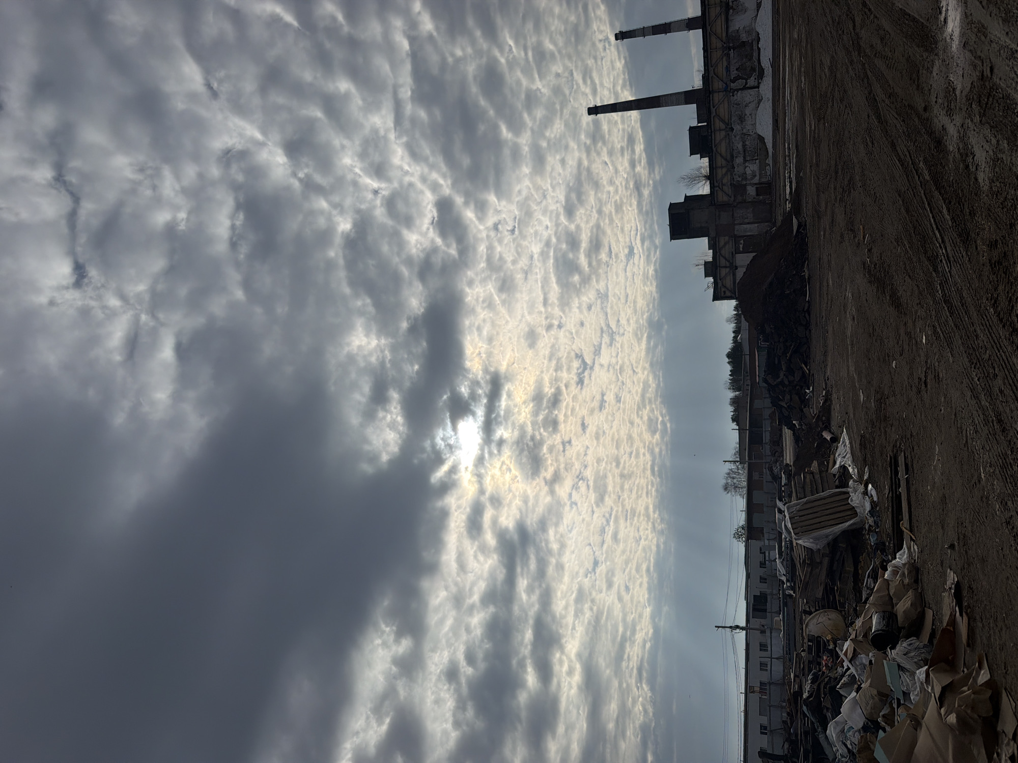 The image depicts a desolate industrial landscape under a dynamic, cloudy sky. The upper two-thirds of the frame are dominated by clouds, ranging from dark, dense formations on the left to lighter, textured altocumulus clouds on the right. The sun is partially obscured by clouds in the central upper portion, creating a bright spot and casting rays that illuminate the surrounding cloud structures.
In the mid-ground, on the right side, several industrial structures are visible. Two prominent, tall chimneys, one made of brick and the other appearing metallic, rise against the sky. Below them are dilapidated factory buildings, exhibiting signs of decay and possibly partial collapse, suggesting an abandoned or disused site. Sparse, bare trees can be seen near these structures.
The foreground and much of the lower portion of the image consist of dark, uneven ground, likely dirt or mud, marked with distinct tire tracks. Along the base of the industrial buildings and extending into the foreground, piles of assorted debris, construction waste, and trash are scattered, reinforcing the sense of neglect and disarray.
There is no specific human activity taking place, and the scene conveys an atmosphere of industrial decay and abandonment, contrasted with the natural beauty of the sun-drenched, cloudy sky. The time of day appears to be daytime, possibly late afternoon or early morning given the light quality. No visible text is discernible within the image.