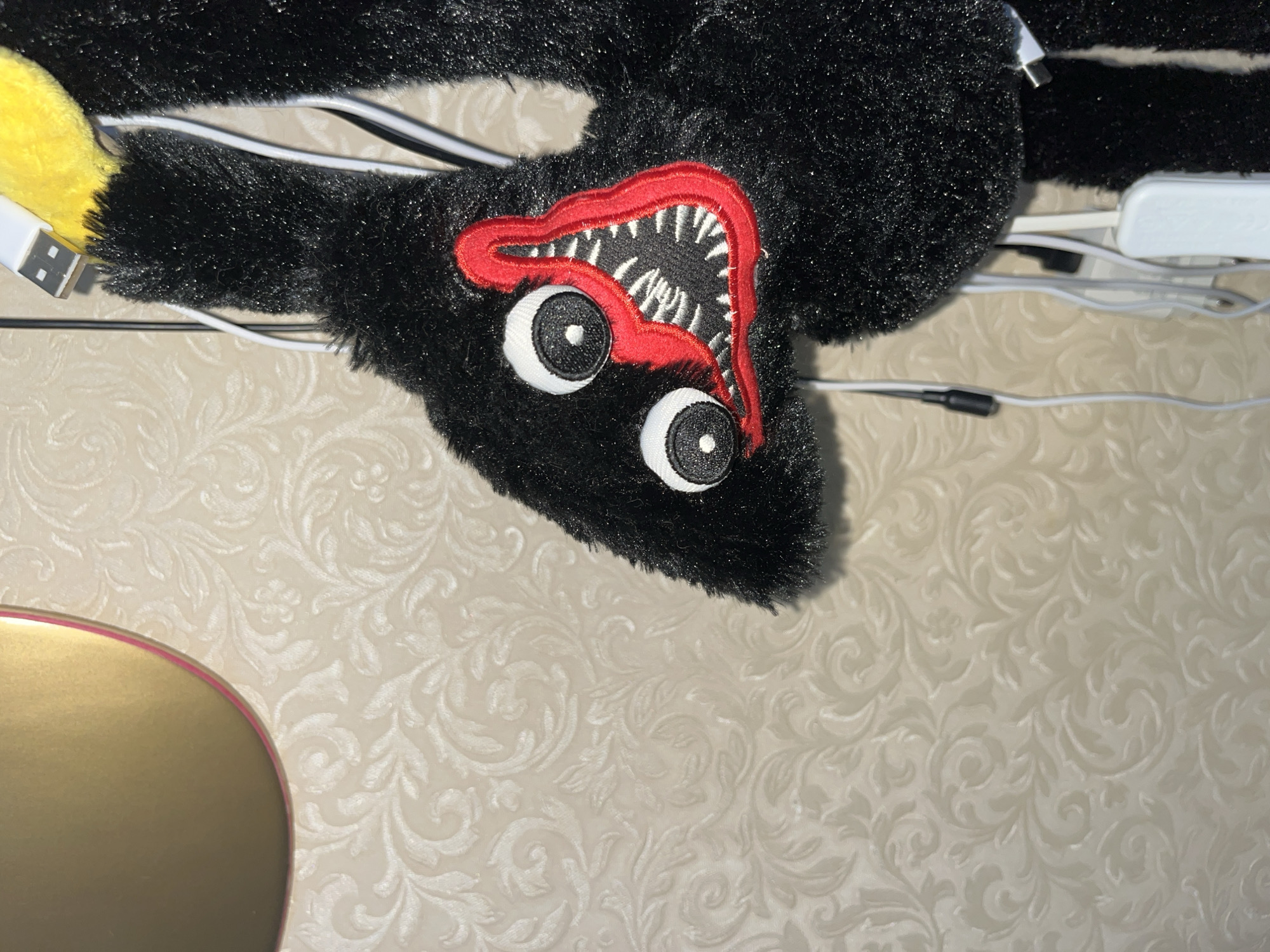 A close-up, eye-level shot shows a black, plush toy with large, white, circular eyes with black pupils. The toy has a red mouth with sharp white teeth. The toy is hanging, and several white cables and a white USB adapter are visible near it, attached to a textured, cream-colored wall with a floral pattern. To the left of the toy, a portion of a gold-colored, circular object with a pink edge is visible.
