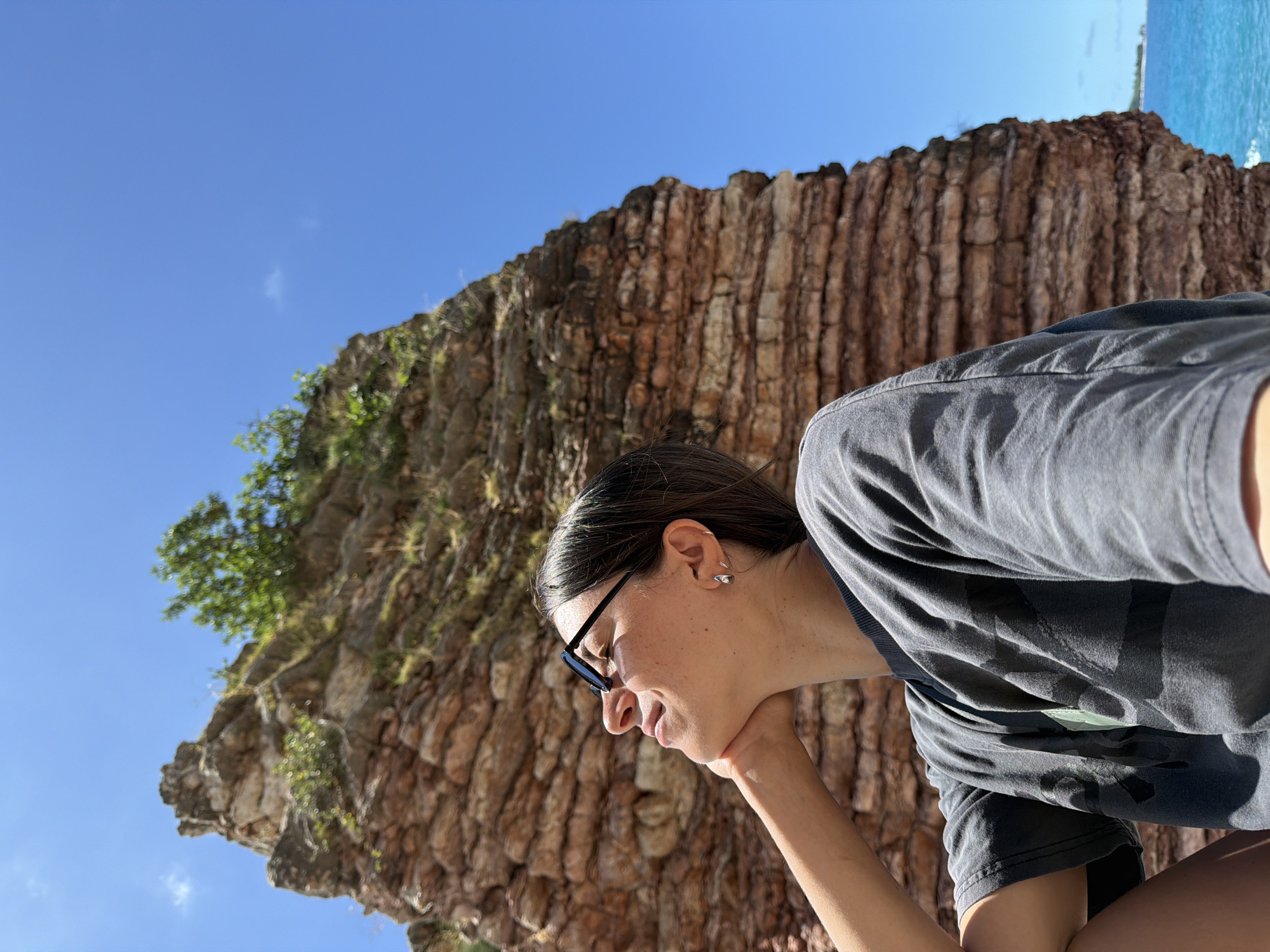 The image presents a sunlit, outdoor scene, likely in Boreti, Montenegro, under a clear blue sky. The main subject is a young woman, captured in profile, looking down and to her left with a calm, slightly smiling or contemplative expression. She has dark hair, wears dark-rimmed glasses, a dark grey t-shirt, and small silver earrings. Her right hand is gently placed under her chin.
Dominating the background is a large, striking reddish-brown rock formation or cliff face. This geological feature displays distinct horizontal layers and striations across its surface, suggesting a unique natural erosion or formation. Green foliage, including small trees and bushes, grows on top and along the sides of the rock, providing a contrast of vibrant life against the rugged stone.
In the upper background, beyond the rock formation, a serene expanse of blue water is visible, indicating the presence of the sea or ocean, with a distant, faint landmass or horizon line. The bright daylight and clear sky suggest it is midday or early afternoon. The overall scene conveys a tranquil coastal environment, possibly from an elevated viewpoint. No visible text is present in the image.