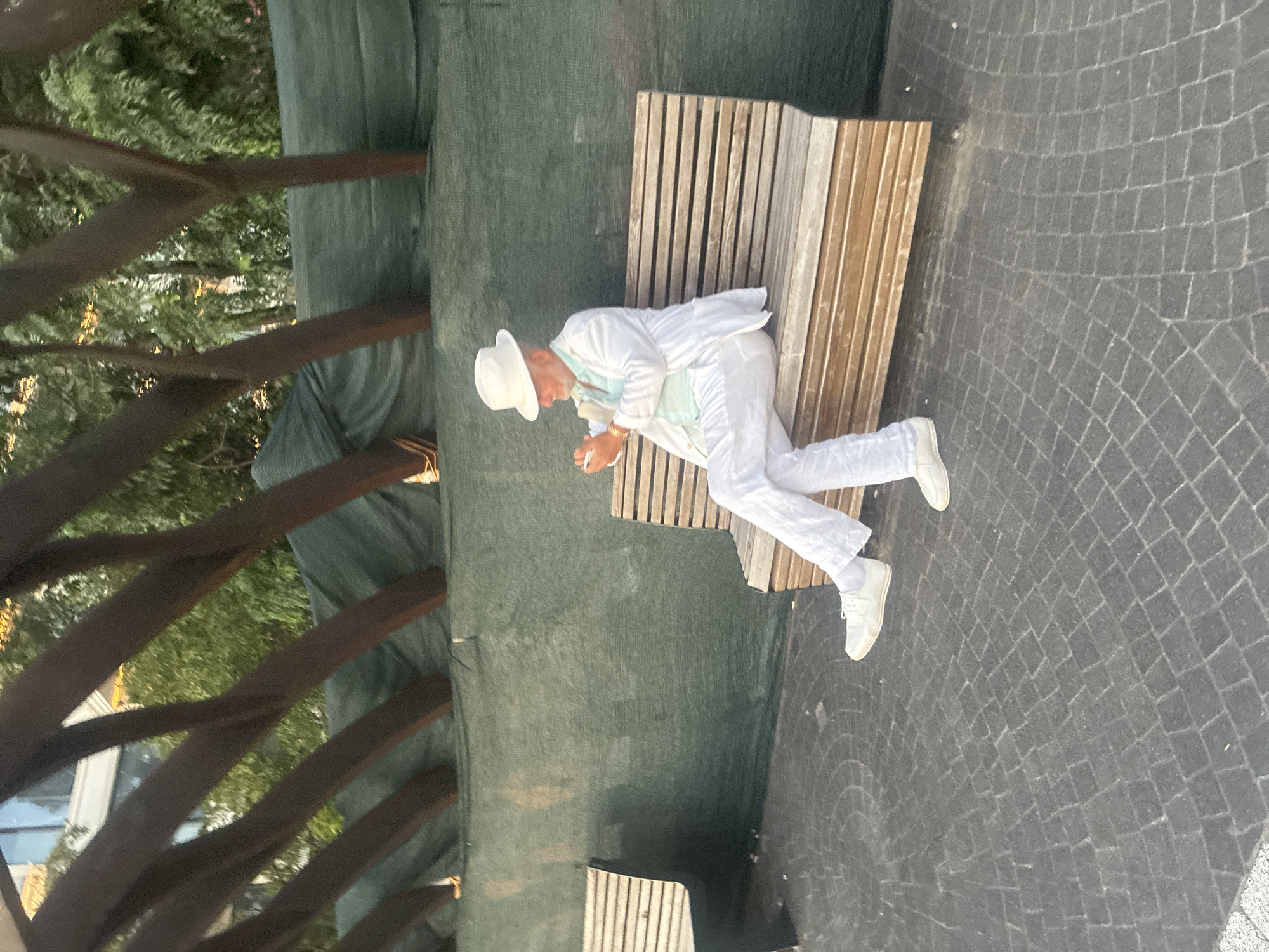 A man in a white suit and white hat is sitting on a wooden bench outdoors, looking at his phone. He is wearing a light blue shirt under his jacket. The bench is next to a dark gray paved area with a circular pattern. Behind the bench is a green mesh screen. Dark wooden beams are visible above the screen, and trees and a building can be seen in the background. The scene appears to be in a public outdoor space in Sochi, Russia.