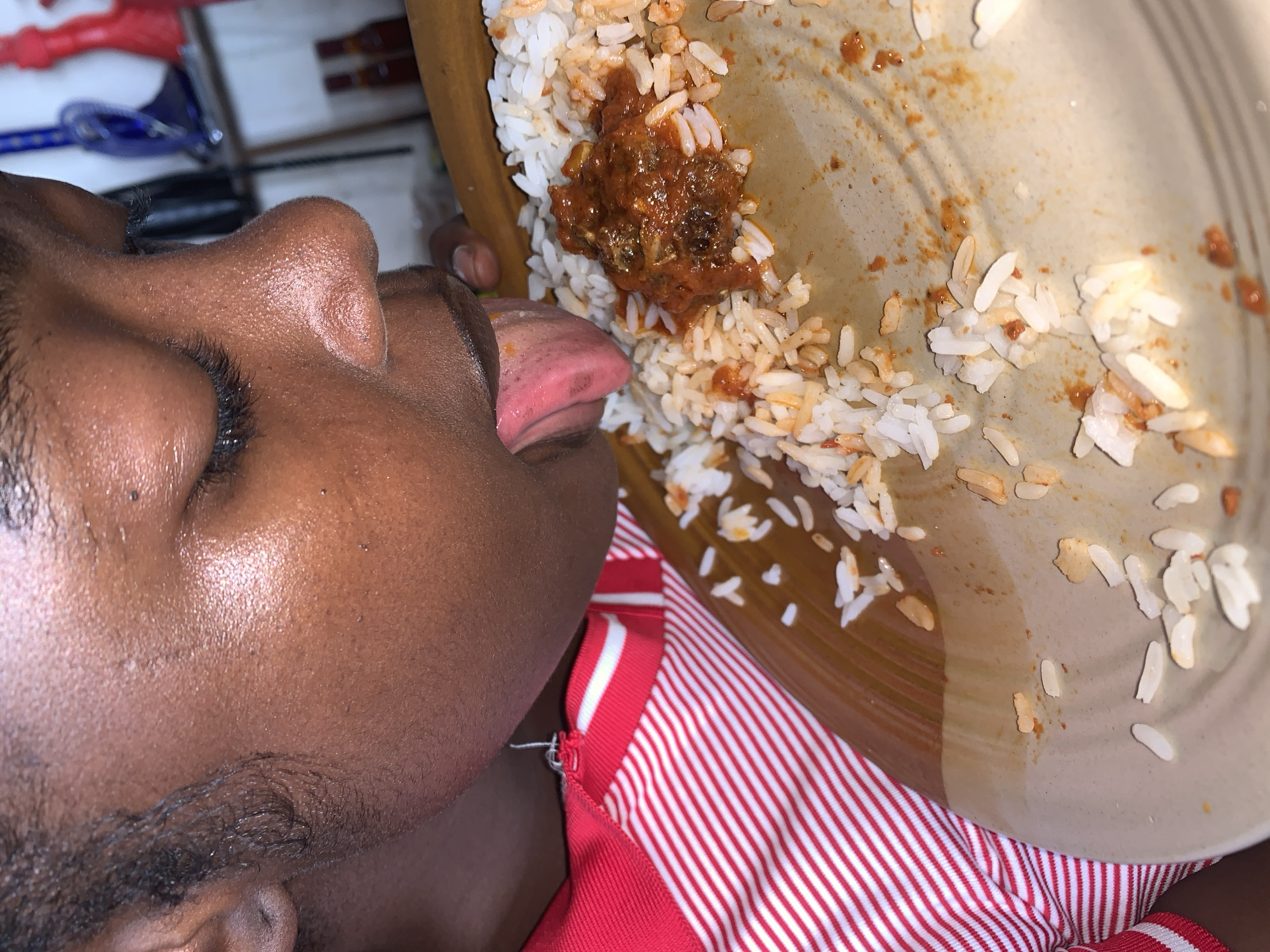 A close-up, tilted image shows a person with dark skin licking a plate of food. The person's face is seen in profile from the right, with their eyes cast downwards or closed. Their tongue is extended, touching a light brown plate that contains remnants of white rice and a rich, reddish-brown stew, which appears to have small pieces of meat or fish. Much of the meal has already been consumed, leaving scattered grains of rice and streaks of sauce on the plate.
The person is wearing a red garment with thin white horizontal stripes, visible on their shoulder and chest. The skin on their face has a subtle sheen, and short, dark hair is visible around the eye and cheek area. The background is blurred, showing indistinct indoor elements that suggest a domestic setting. The activity portrays a thorough enjoyment of the meal, as the person is seen licking the plate clean. There are no specific indicators for the time of day or weather within the image itself. No text is visible in the media. This scene takes place in the context of New Stadium, Nigeria.