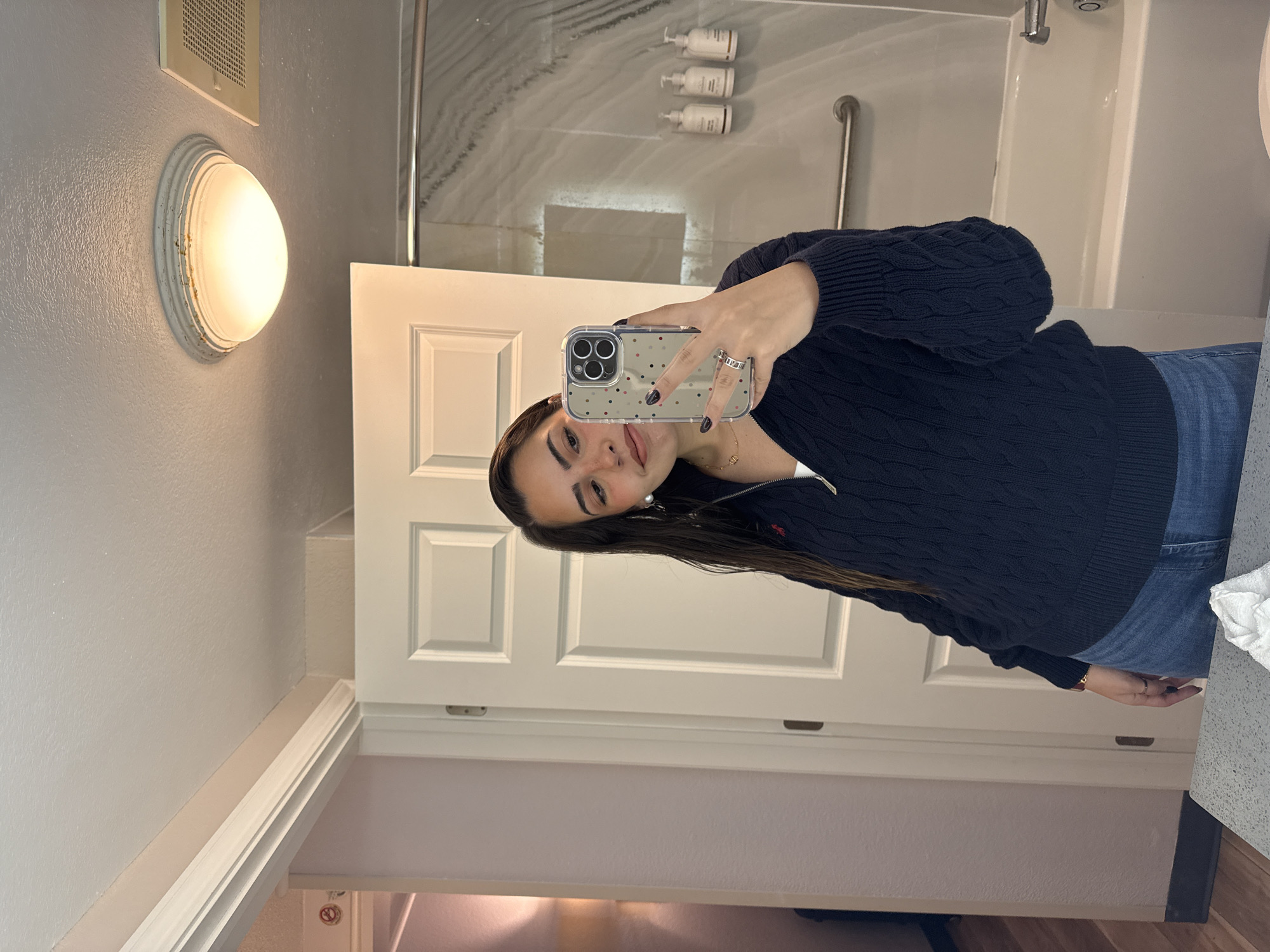 A young woman is taking a selfie in a bathroom. The bathroom appears to be in a hotel. The woman is wearing a dark blue cable knit sweater and jeans. She is holding a smartphone with a clear case with colored dots. The bathroom has a white door, light gray walls, a round light fixture above the door, and a bathtub/shower with a marble-like surround. There are toiletries visible in the shower area. I cannot determine the exact location in San Antonio, United States of America.