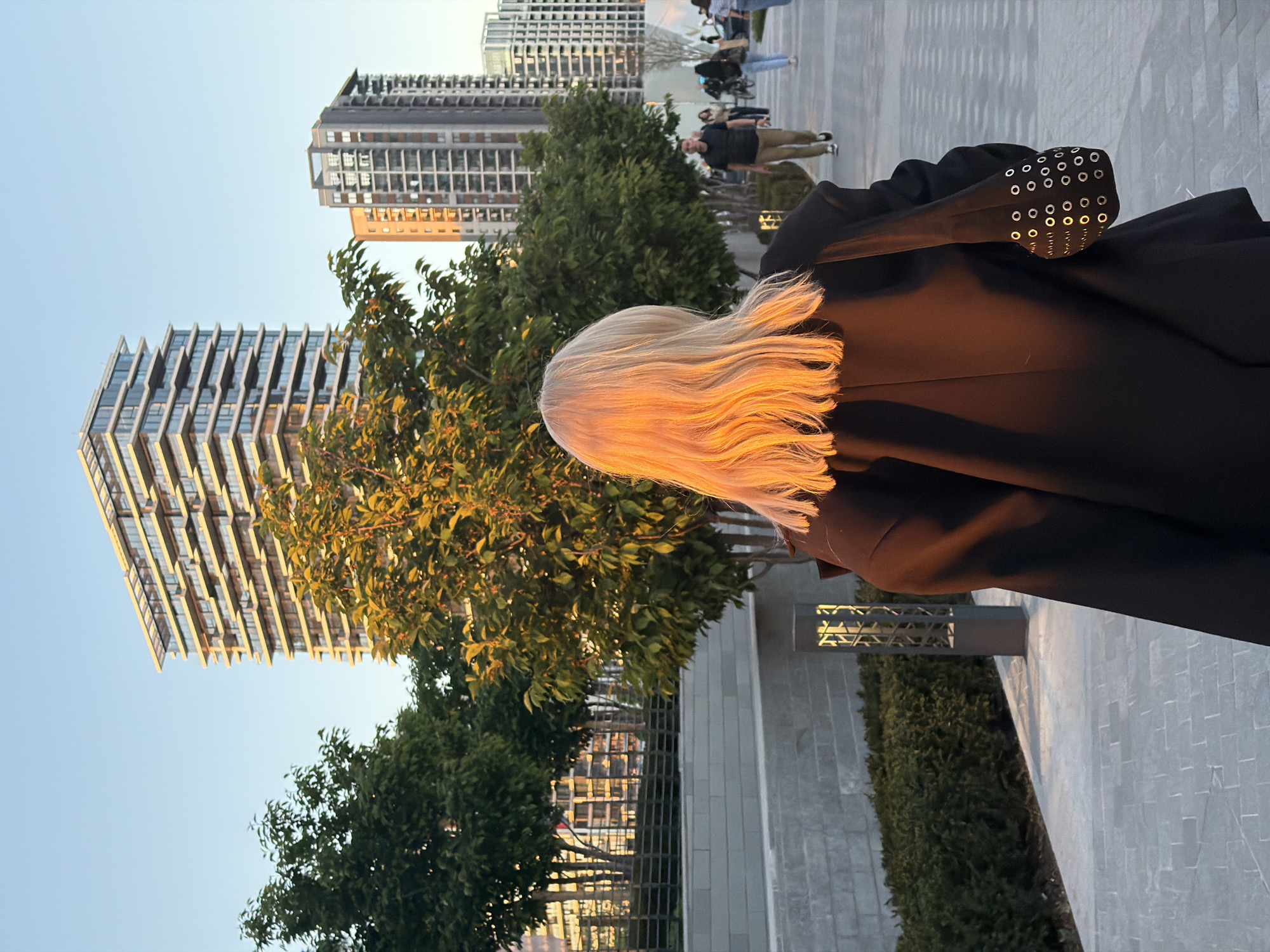 The image depicts an urban outdoor scene in Belgrade, Serbia, during the golden hour of late afternoon or early evening. The primary subject is a person viewed from behind, whose long, flowing blonde hair is strikingly illuminated by the warm, golden sunlight. They are wearing a dark, likely black, jacket, which features a distinctive design element on the left sleeve: multiple rows of small, circular silver eyelets or grommets.
In the background, several modern high-rise buildings dominate the skyline, characterized by their glass facades and unique architectural structures. The sky above is clear and light blue, indicating fair weather. The urban landscape includes green trees and bushes, providing natural elements amidst the structures. A paved pedestrian walkway, constructed from light-toned bricks or tiles, extends through the scene.
Further in the background, other individuals are visible, walking casually along the promenade, suggesting a public and active area. A modern street lamp with a geometric patterned light fixture is partially visible in the bottom right corner. No visible text is present in the image. The scene conveys a sense of a vibrant, contemporary city bathed in the soft, glowing light of sunset.