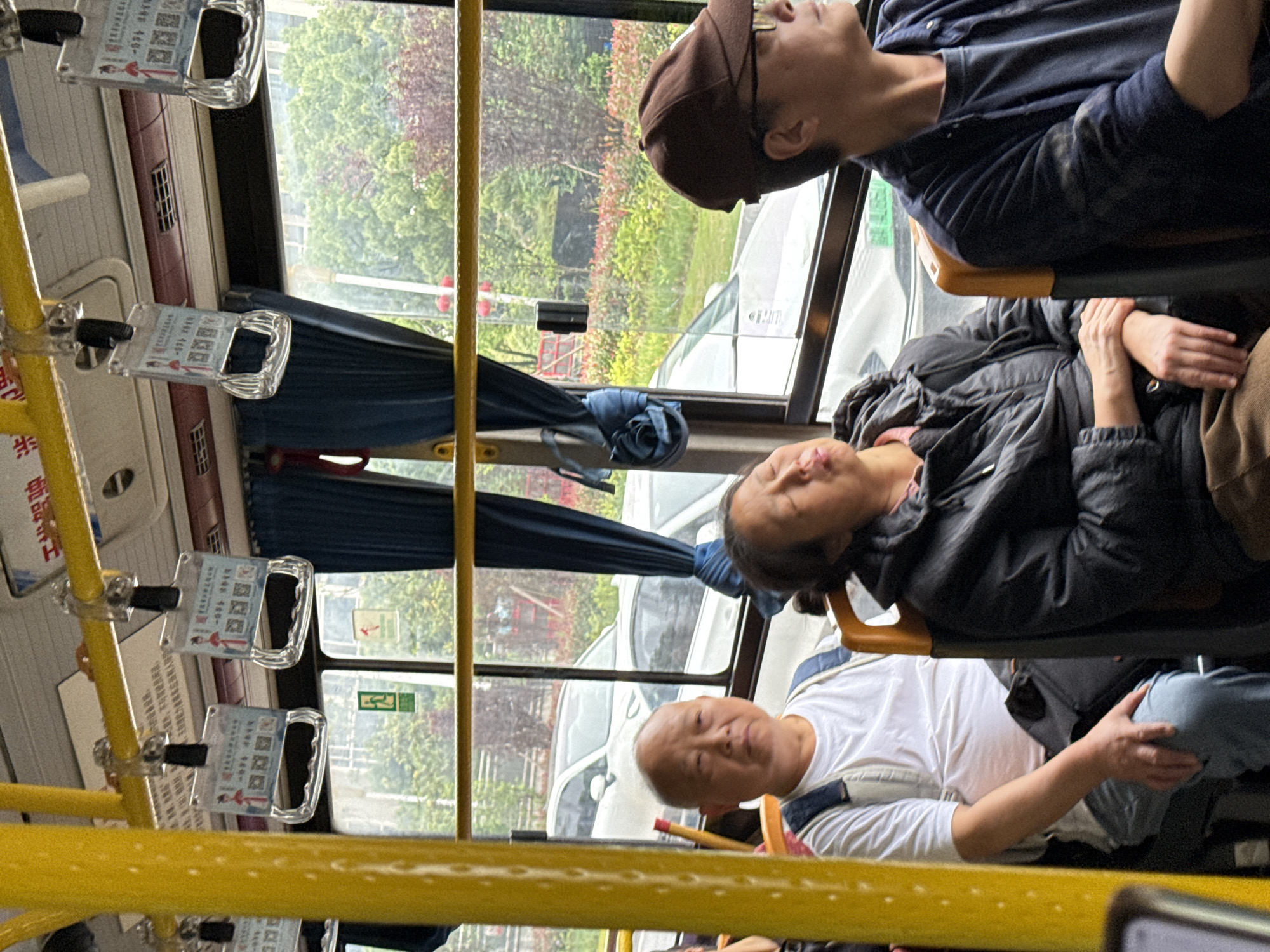 The image shows the interior of a bus in Jiangjin, China. Three individuals are visible, all seated. The person at the front wears a white shirt and jeans. The second individual wears a dark jacket and brown pants, seated behind the first. A third individual, wearing a dark blue shirt, is seated behind and to the right of the second person. All three seem to be resting or sleeping. Through the bus windows, trees, vehicles, and buildings are visible. Yellow handrails and fare information are attached to the bus ceiling.