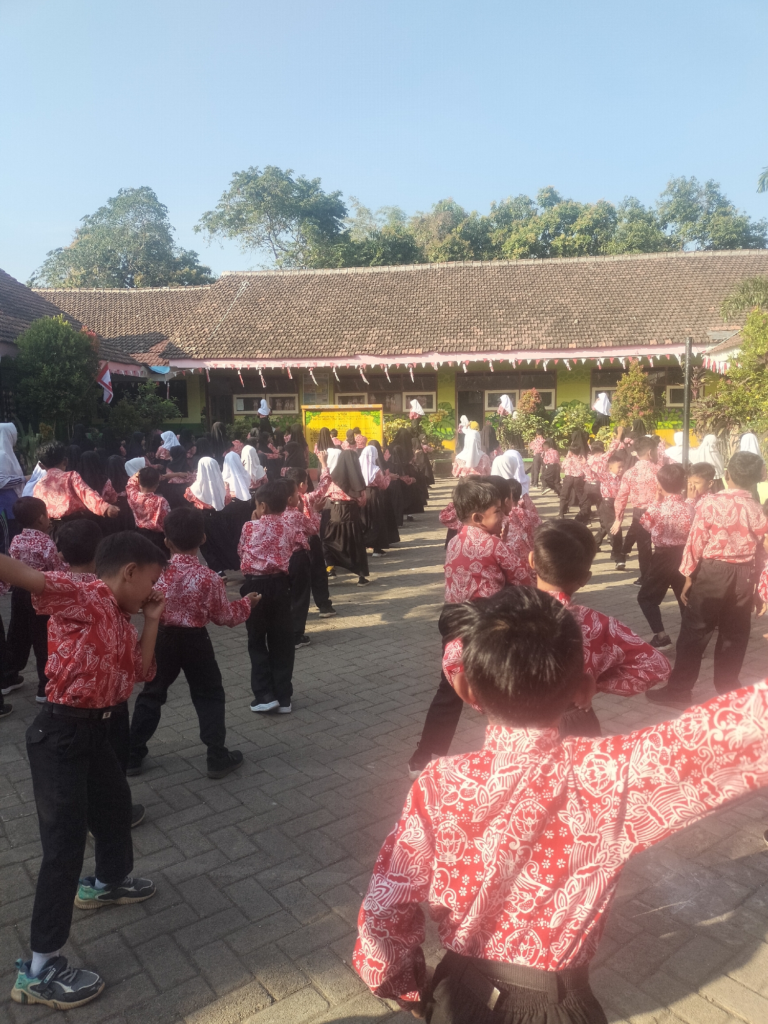 In Gunung Tugel, Indonesia, a group of young students, predominantly male and some female, are engaged in an outdoor activity in what appears to be a schoolyard. The children are dressed in matching red and white batik shirts and black pants, with some girls wearing headscarves. They are arranged in a semi-circle, facing a building with a red-tiled roof and green walls, which is decorated with white and red bunting. The ground is paved with interlocking grey bricks. The students seem to be participating in a synchronized exercise or dance, with many of them holding their arms out or in motion. The scene is set under a clear blue sky with lush green trees visible in the background.