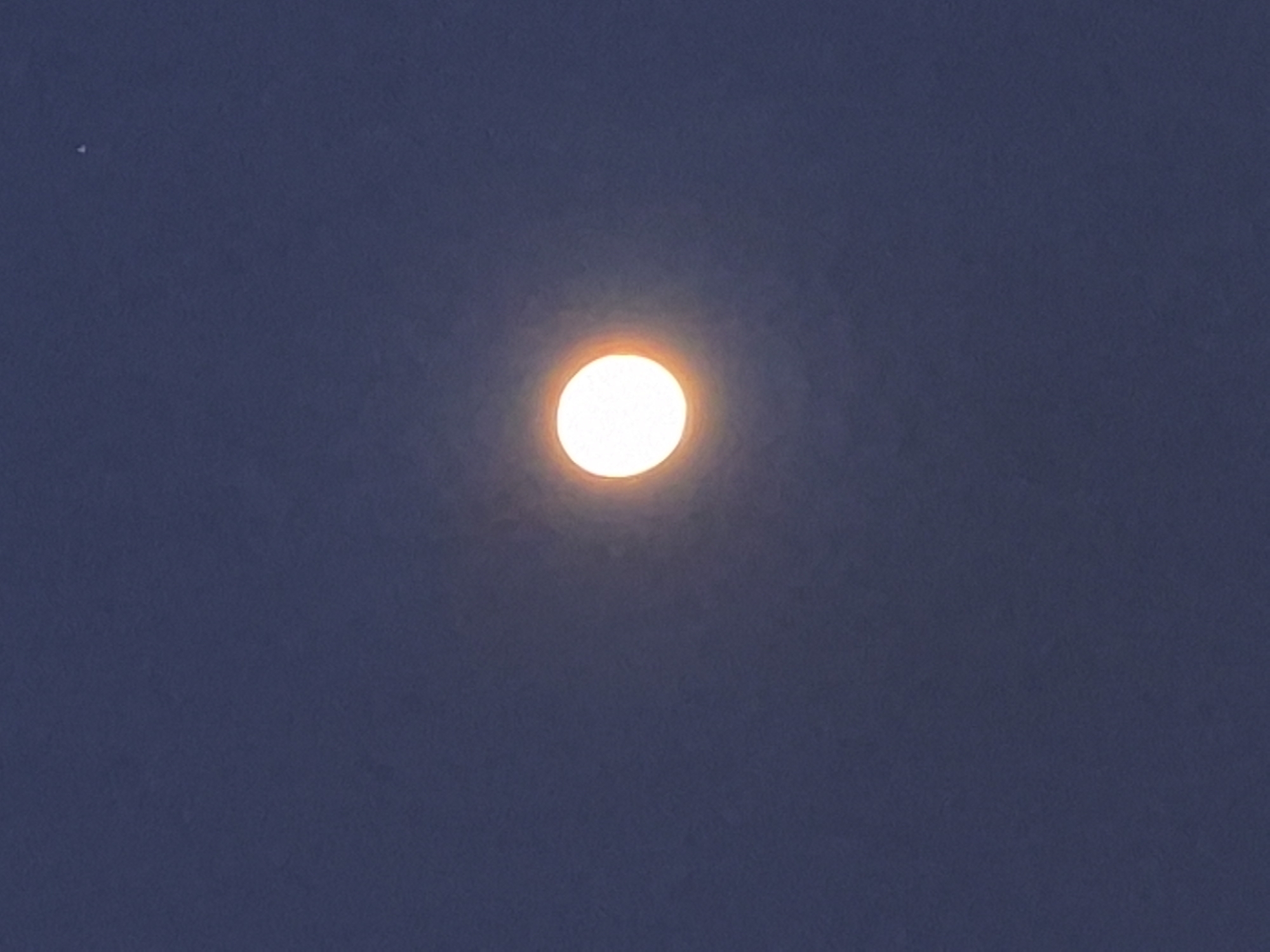 The image shows a full moon in a dark blue sky. The moon is bright and round, with a slight orange glow around its edges. There is a small, bright point of light, possibly a star, in the upper right corner of the image. The sky is clear and there are no clouds visible. The image is taken from a low angle, looking up at the moon. The overall impression is one of peacefulness and tranquility.