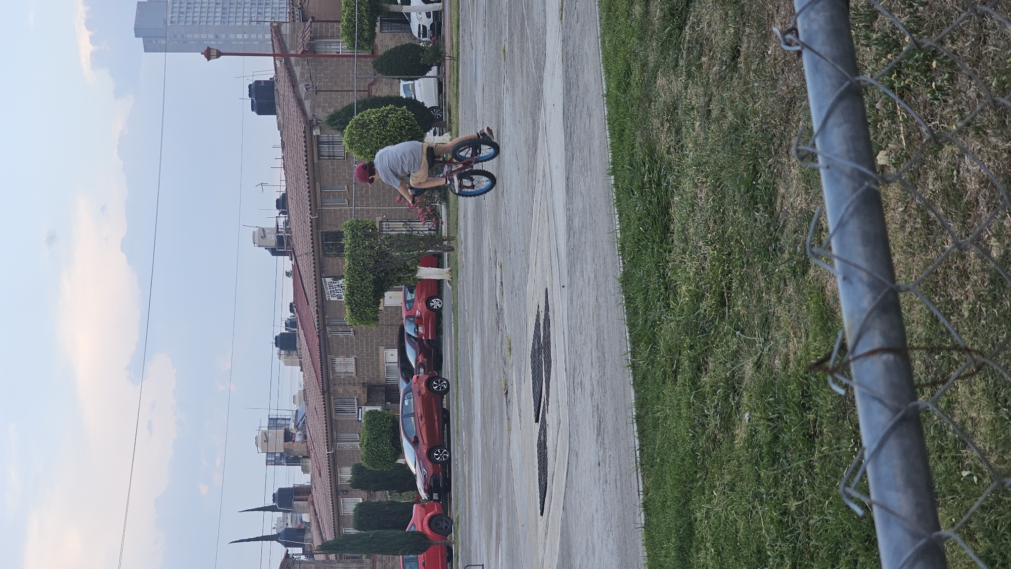 A man on a bicycle is performing a trick on a paved path in San Bernardino Tlaxcalancingo, Mexico. The man is wearing a red hat, a gray shirt, and khaki shorts. He is positioned on the bike with both wheels off the ground. In the background is a row of brick apartment buildings with water tanks on the roofs. Red cars are parked in front of the buildings. There are manicured shrubs and trees alongside the buildings and path. A chain-link fence runs along the right edge of the image, separating the path from a grassy area. In the distant background, a tall building is visible under a cloudy sky. Dark tire marks are on the paved path.