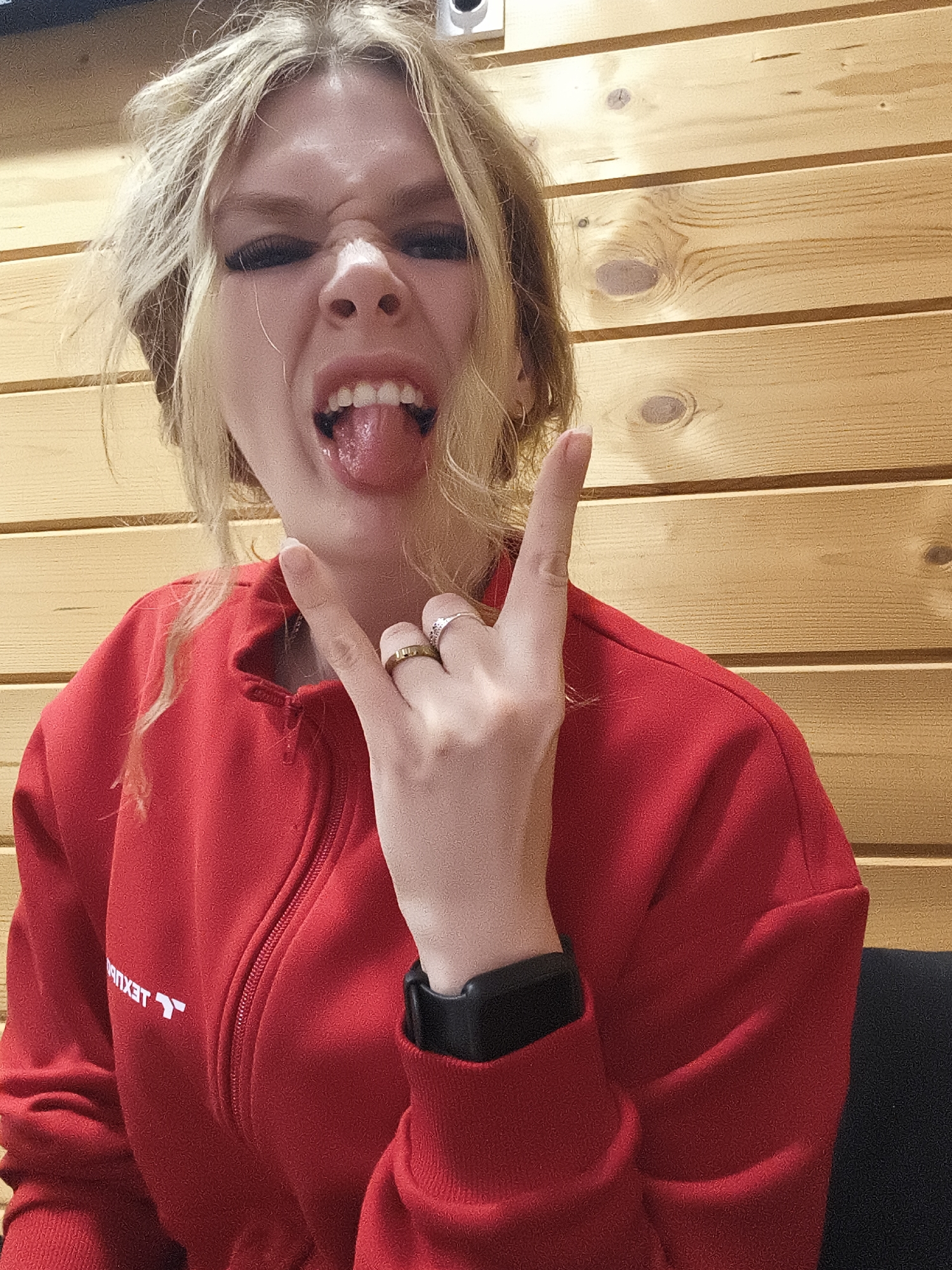 A blonde woman is indoors, posing in front of a light wood paneled wall. She is wearing a red jacket with a zipper and white text on the left chest. She is sticking her tongue out and making a "rock on" hand gesture. She wears a black smartwatch on her left wrist and rings on her fingers. The location is Русский Вожой, Russia.