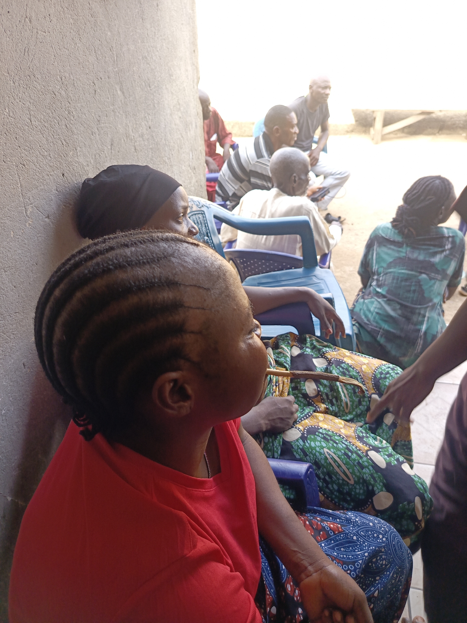 In Akwanga, Nigeria, a group of people are sitting on blue plastic chairs. A woman in the foreground wears a red shirt and has braided hair. Beside her, a woman with a black head covering leans against a wall. Behind them, several people are seated, including men in striped shirts and a woman in a green and blue patterned dress. A wooden stick rests on the lap of the woman in red. The scene appears to be outdoors, next to a light-colored wall.