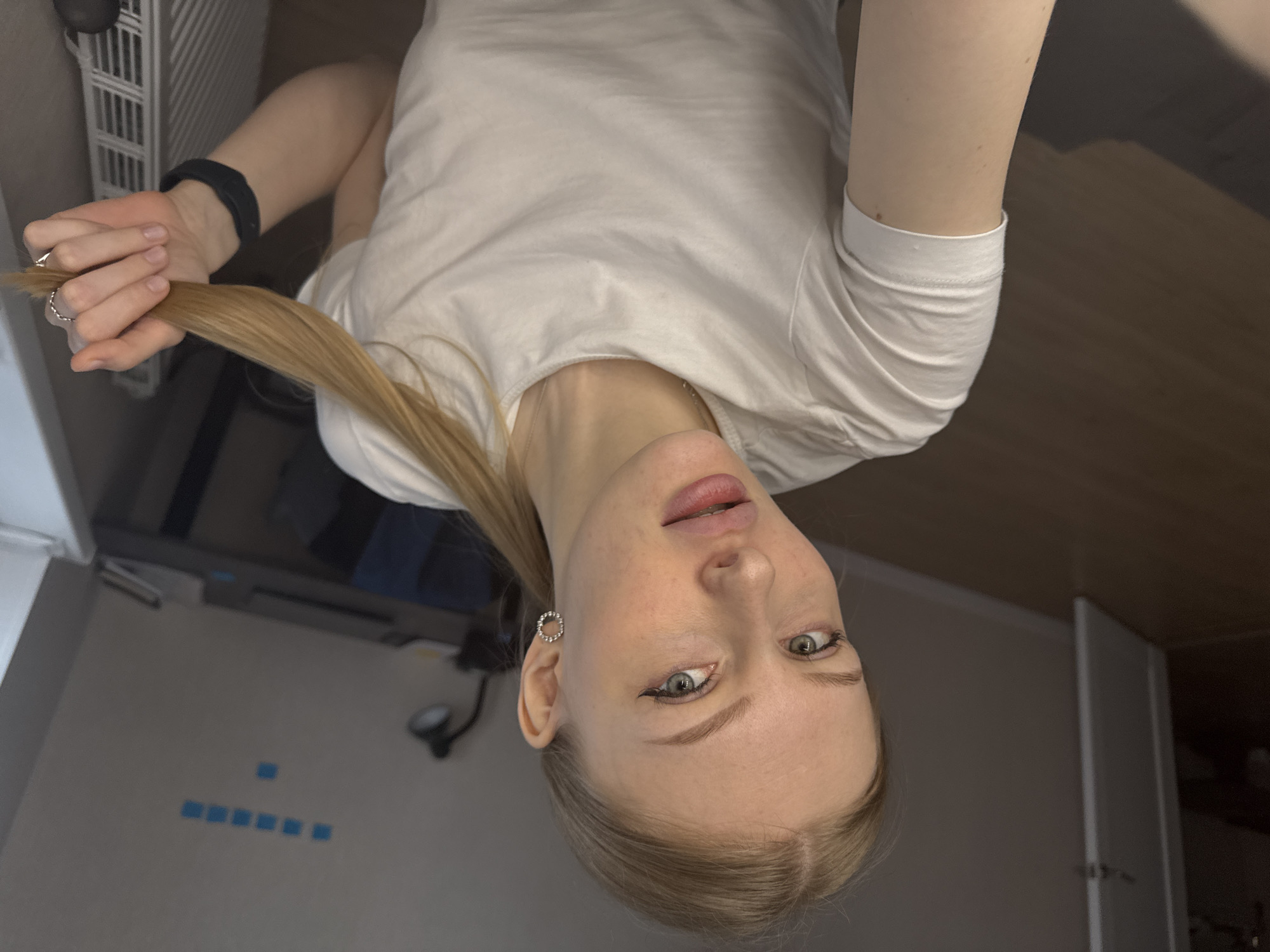 The media file captures a close-up indoor portrait of a young woman, presented with a 90-degree clockwise rotation. The woman, with light blonde or light brown hair, is looking directly at the viewer. Her eyes are green, and she has subtle eyeliner and slightly parted lips, showing some teeth. She wears a simple white crew-neck t-shirt and circular, possibly diamante, earrings. Her left hand is visible, holding a section of her hair, and a ring is present on her left ring finger. A black wristband is visible on her right arm.
The background reveals parts of a room. On the left side, a white radiator or vent is partially visible against a light gray wall. Further back on the wall, a series of small blue square stickers are arranged in a horizontal line, with one isolated sticker above them. Other background elements are darker and out of focus, including what appears to be part of a dark furniture item or doorway on the right. The lighting is even and soft, suggesting an indoor setting with artificial illumination. The image does not contain any visible text. The location context for this scene is Perm, Russia.