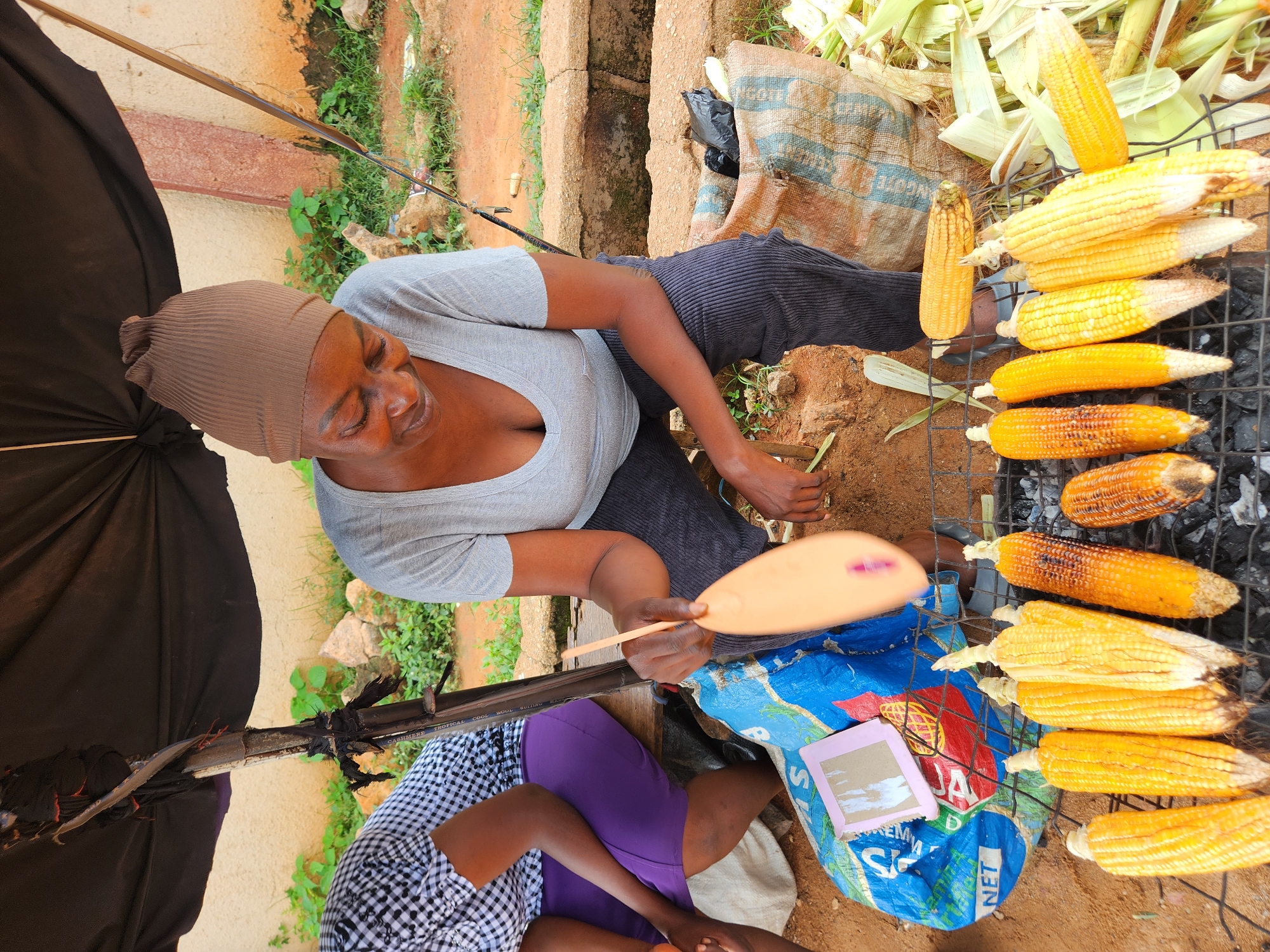 A woman, wearing a brown headwrap and a grey t-shirt, is grilling corn in an outdoor market in Sabon Tasha, Nigeria. She is holding a fan made of a light-colored material, and appears to be tending to the corn. Several ears of corn are arranged on a metal grill over hot coals. Another woman, wearing a black and white checkered shirt and a purple skirt, is seated to the left of the main subject. She is partially obscured by a large blue plastic bag. A large burlap sack filled with corn husks and a black plastic bag is visible in the background. The scene is set against a backdrop of a wall and some vegetation. The ground is dirt.