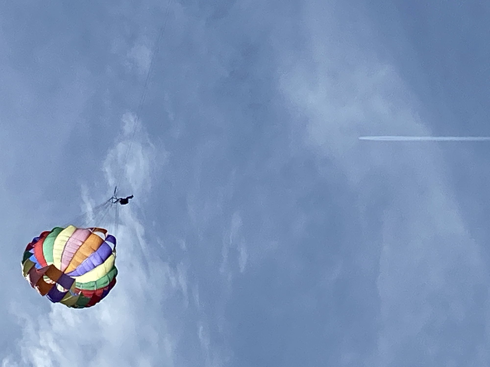 The image shows a parasailor suspended beneath a colorful parachute against a blue sky with clouds. A contrail from an airplane is visible in the distance. The location is not discernible from the visual data. There is no indication that this scene is in Prijevor, Montenegro.