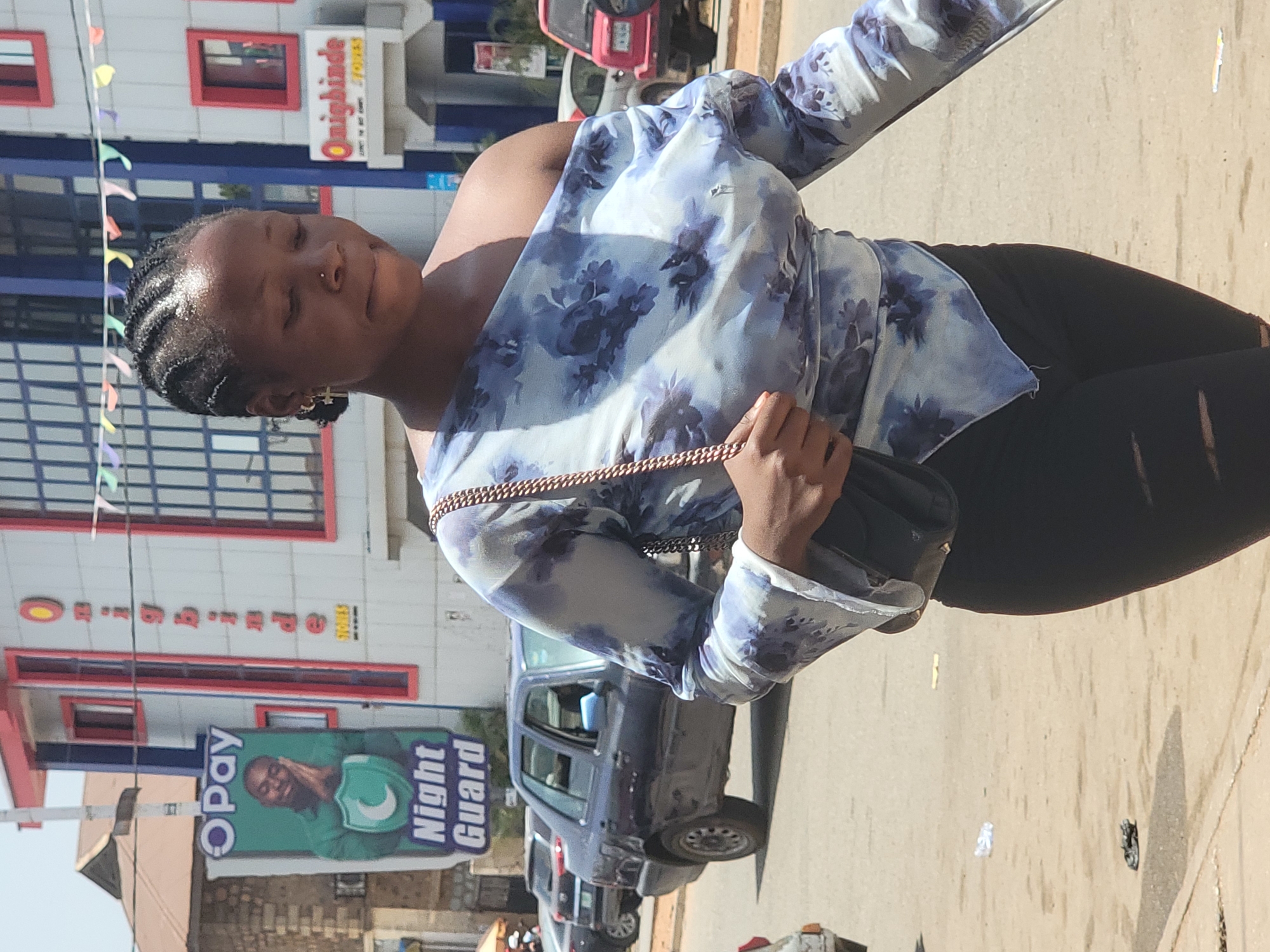 A woman walks along a paved street in Jos, Nigeria, under bright daylight. She is centrally positioned, seen from the waist up, with her body angled slightly away from the camera. She wears a light blue and white floral-patterned, long-sleeved top and dark-colored pants, and carries a small, dark handbag with a gold chain strap. Her hair is braided close to her head, and she has a small nose piercing. Her eyes are closed or cast downwards, and a subtle smile plays on her face, suggesting a relaxed or contemplative mood.
The background features commercial buildings lining the street. One building is light-colored with red-framed windows, and a partially visible sign reads "Onigbinde Stores." In the mid-ground, an "Opay Night Guard" billboard prominently displays a smiling man and the Opay logo. Several cars are parked or driving on the street, and colorful string flags are visible across a space between buildings in the distance. Strong shadows are cast on the ground, indicating a sunny day. The scene captures a typical urban environment in a Nigerian city.