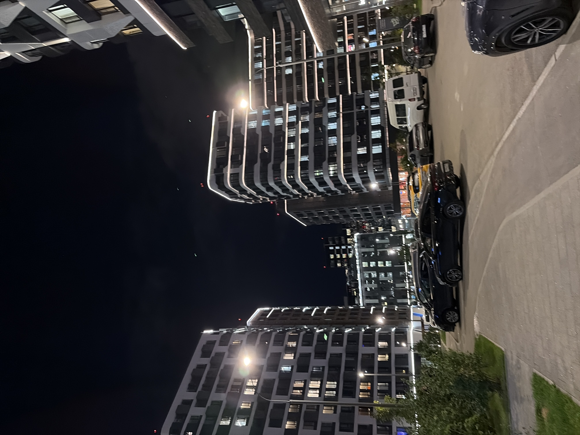 This image depicts an outdoor nighttime scene in a modern urban environment. In the foreground, a paved walkway leads to a parking area where several vehicles are parked, including black sedans, a silver sedan, and a white van. Behind the parking lot, a series of multi-story buildings with illuminated windows and architectural lighting are visible. The sky is dark, suggesting it is nighttime. The overall impression is of a residential or mixed-use development. Based on visual cues such as architectural style and vehicle models, this scene is likely located in Moscow, Russia.