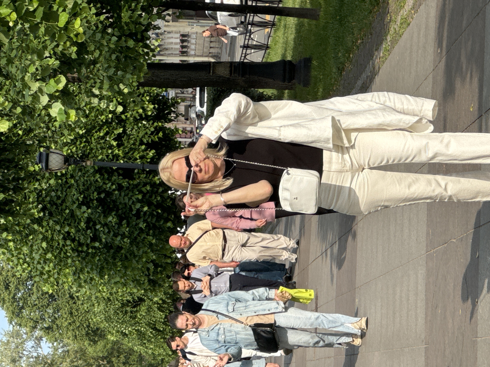 The image depicts a woman in the foreground walking along a paved sidewalk lined with trees and a lamppost, characteristic of a park or boulevard in Saint Petersburg, Russia. She is wearing sunglasses, a black top, a white jacket draped over her shoulders, and light-colored pants. A white crossbody bag is visible. In the background, a procession of people walks in the same direction, some engaged in conversation. Further in the distance, a woman is seen walking on the grass beside a street with parked cars. The scene is illuminated by sunlight, casting shadows on the sidewalk.