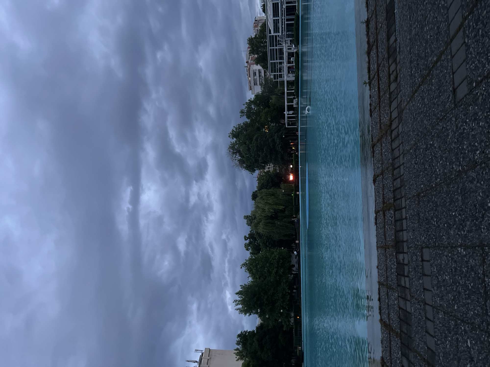 The image depicts a long, rectangular body of water, possibly a decorative pool or artificial canal, situated in an urban area in Plovdiv, Bulgaria, under a cloudy, overcast sky. The water's surface is relatively still, with slight ripples. To the right of the water is a paved walkway composed of grey bricks or stones. Beyond the body of water, a line of trees separates the water from buildings, some of which have a white, modern architectural style. Some trees and vegetation are visible further back. There are no people discernible in the frame. The lighting suggests either late evening or a heavily overcast day. The overall mood is subdued and somewhat melancholic due to the dark sky and muted colors.