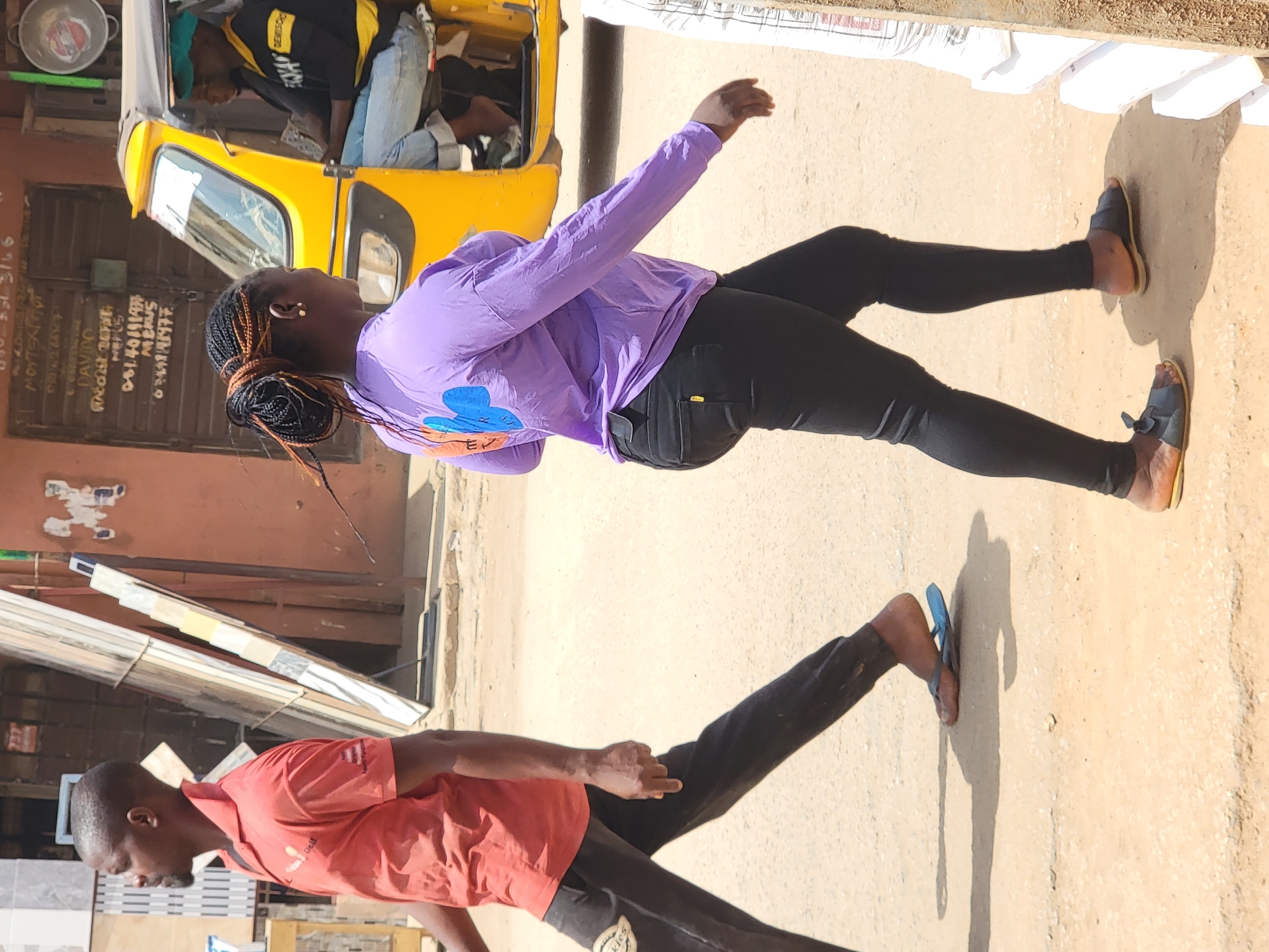 The image shows an outdoor scene in Maiburiji, Nigeria, with two people walking on a street. A woman in a purple long-sleeved shirt and black leggings walks with her arms raised slightly. Nearby, a man wearing a red t-shirt and dark pants is also walking. In the background, there is a yellow auto rickshaw, a building with signs, and some building materials. The ground appears to be a sandy color.