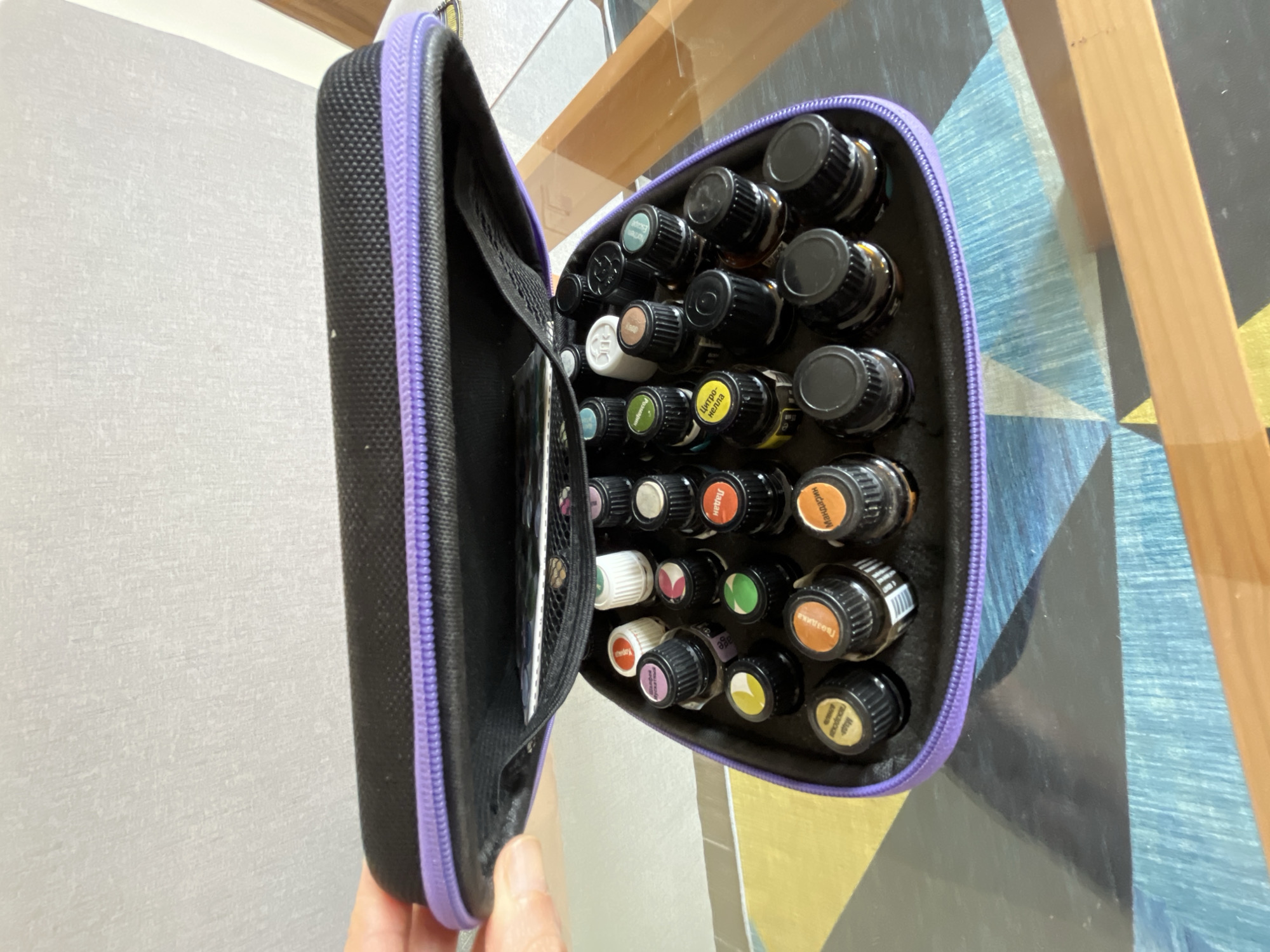 In the city of Xuân Thành, Vietnam, a black and purple zippered case is open, revealing an assortment of small glass bottles with black caps, likely essential oils. The case is positioned on a patterned surface composed of blue, yellow, and black geometric shapes, with a wooden surface visible in the foreground. A hand, with visible fingernails, is partially seen on the left side of the image, holding the case open. The background consists of a textured, light-colored wall.
