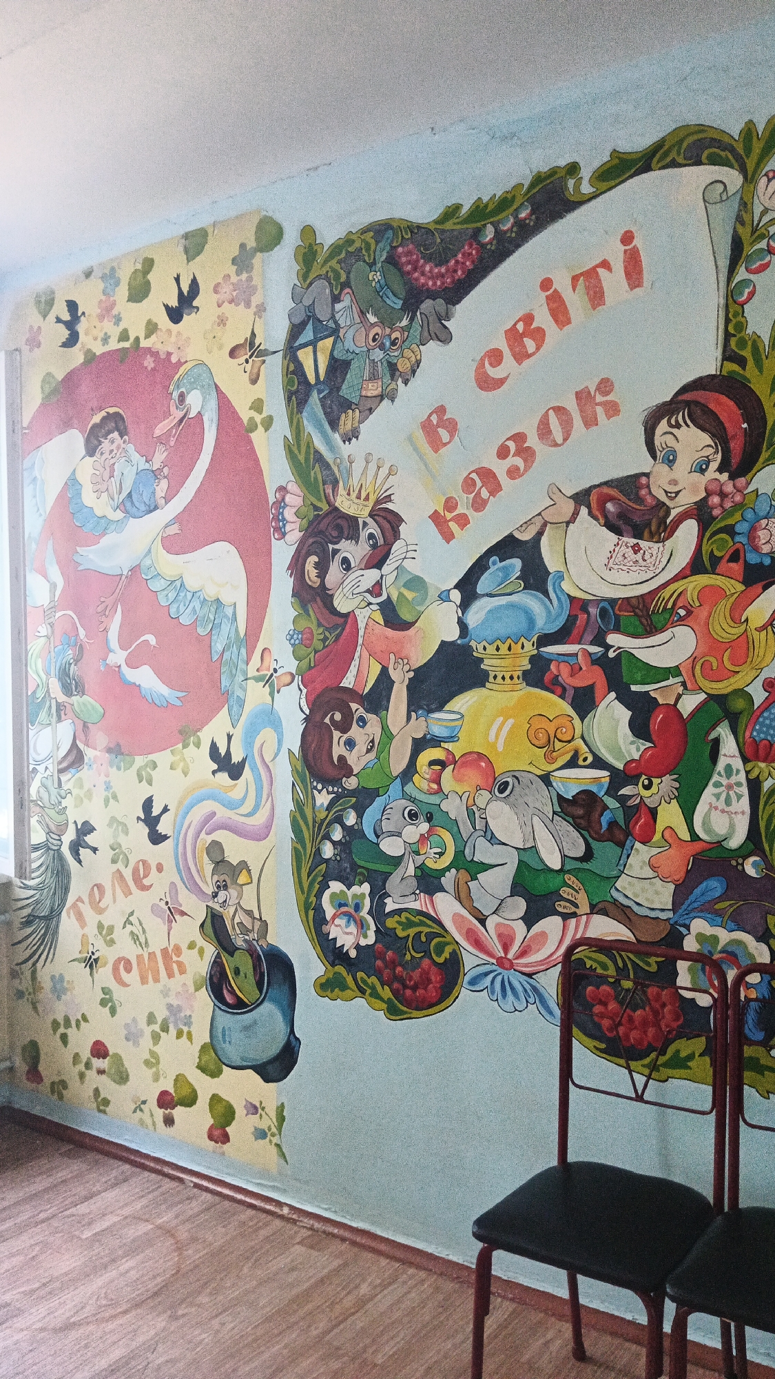 This image depicts a colorful mural on a wall in what appears to be a children's room or a public space like a school or kindergarten, likely in the city of Kryvyi Rih, Ukraine, given the context. The mural is divided into two distinct sections.
The left section features a light yellow background with a whimsical pattern of small flowers and mushrooms. In the upper left, a child sits on the back of a white swan that has a pink outline. Two smaller white birds are flying near the swan. Below this, there are silhouettes of black birds and colorful abstract wavy lines. Text in Cyrillic letters, reading "теле." and "сик", is visible in this section. A small mouse character is depicted sitting in a blue pot.
The right section of the mural is on a light blue background and displays a border decorated with green leaves and red berries, reminiscent of traditional Ukrainian folk art. This section is a lively scene filled with cartoonish characters. A scroll, also framed by the decorative border, contains Cyrillic text in red letters that reads "В світі казок" (In the world of fairy tales). Prominently featured are a crowned lion, a girl in traditional clothing holding a teacup, a fox, a rooster, rabbits, and a frog, all interacting in a festive, tea-party-like setting around a large yellow samovar.
The lower part of the image shows the floor, which is a wooden parquet with a visible brown baseboard along the wall. Two simple black chairs with red metal frames are placed in front of the mural. The lighting suggests an indoor environment with light coming from the left, possibly from a window. The wall shows some signs of wear and tear, with minor discoloration and scuff marks.