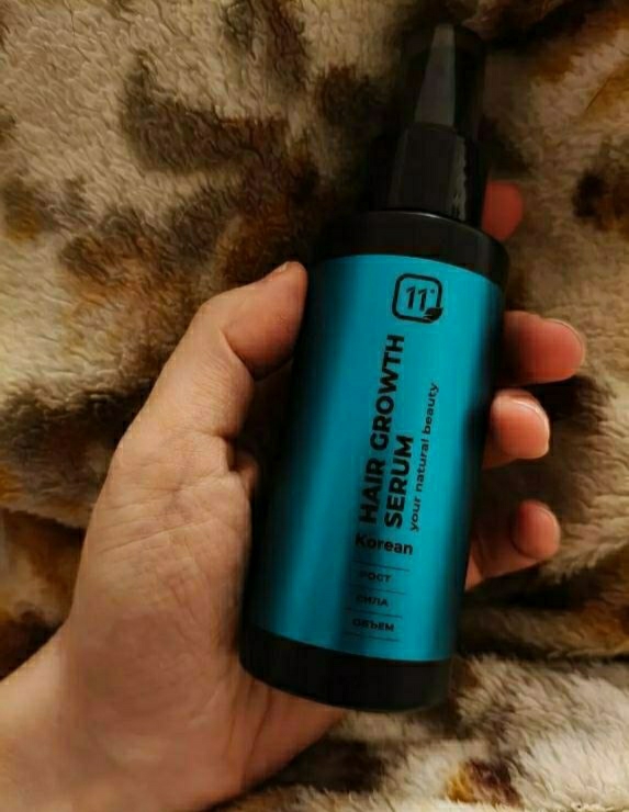 A hand holds a bottle of blue-colored hair growth serum. The bottle has black accents and text in both English and a Cyrillic alphabet. The backdrop is a textured surface reminiscent of brown and tan fur. The location cannot be determined as Minsk, Belarus based on the image.