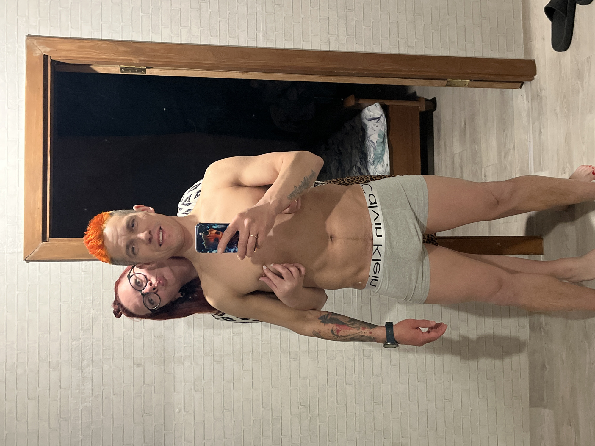 A horizontally oriented medium shot depicts a shirtless, light-skinned man in gray Calvin Klein underwear taking a selfie with a light-skinned woman in eyeglasses. The man has short, bright orange hair and is holding a cell phone. The woman, who has long auburn hair, is positioned lower than the man and has her arm around him. They are standing in front of a dark mirror that is framed with wood. The background consists of white brick-patterned wallpaper and a wooden floor with a pair of black slide sandals near the top right corner.
