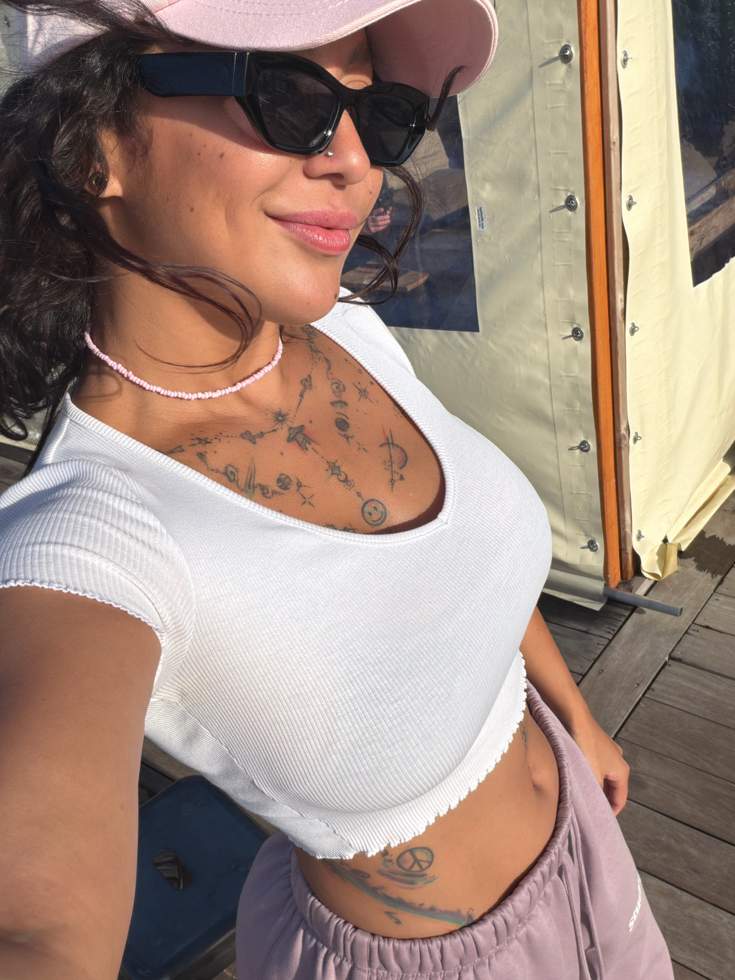 An outdoor, close-up, eye-level, medium shot shows a woman standing in Sangatte, France. The woman is wearing a pink baseball cap, black sunglasses, and a white, cropped, ribbed t-shirt. A thin, pink beaded necklace is around her neck. Tattoos are visible on her chest and stomach, including a peace sign and a smiley face. She is wearing light purple sweatpants. The woman is smiling and looking to her right. She is standing on a wooden deck. Behind her, a pale yellow tent is visible with wooden framing. The scene is lit by bright sunlight, casting shadows.