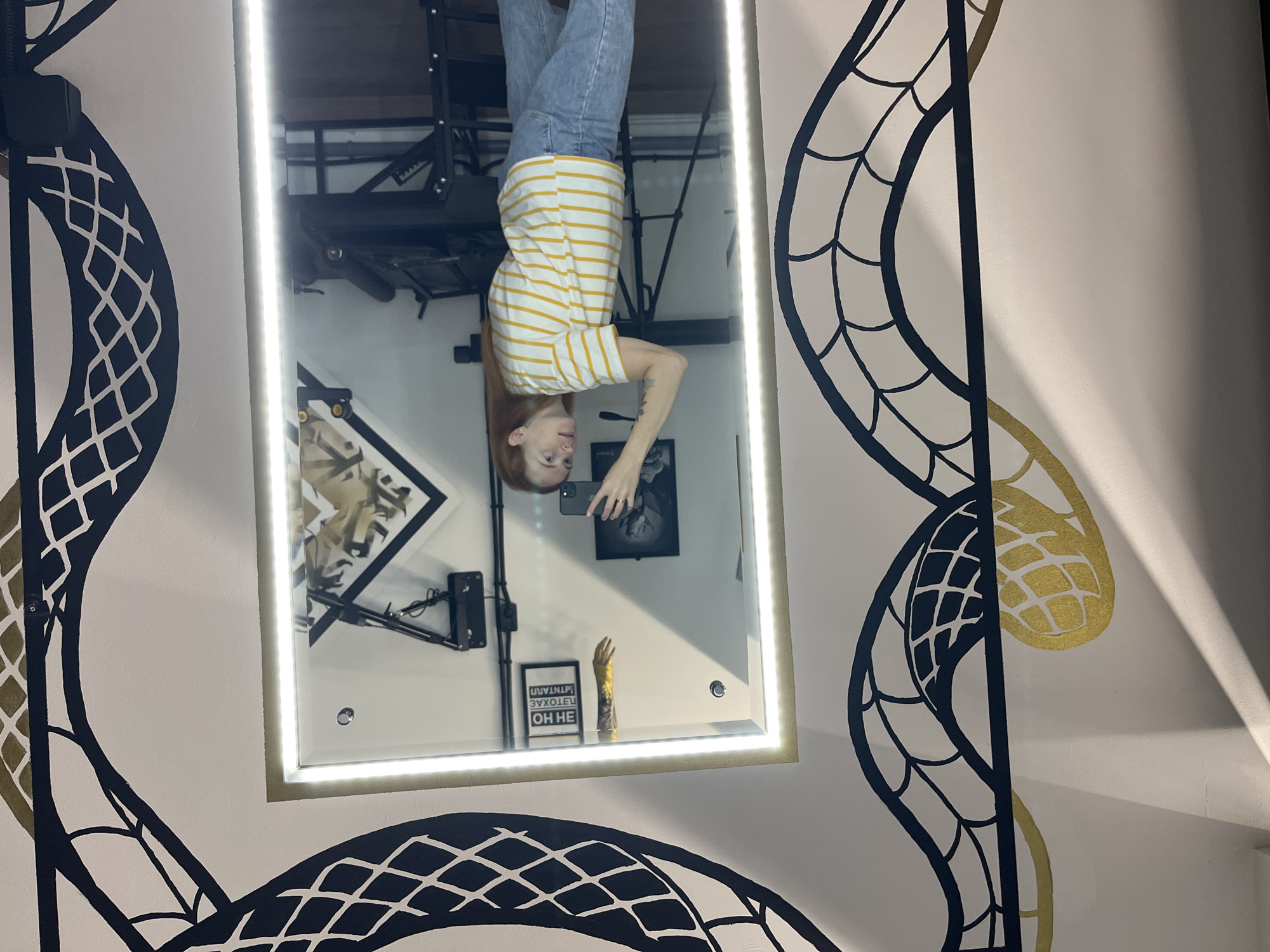 A young woman with red hair, wearing a striped t-shirt and jeans, stands in front of a brightly lit mirror, holding a smartphone to take a selfie. She is positioned within a room decorated with a large, stylized snake mural. The mirror reflects the room behind her, revealing a professional setup with a tripod, a black easel, and what appears to be a tattoo table or similar equipment. A small sign with text in Cyrillic hangs on the wall in the mirror's reflection. The overall environment suggests a studio or personal creative space. The snake mural is partially visible on the left and right sides of the image, wrapping around the mirror. The lighting is bright, with LED strips around the mirror illuminating the scene. The location is within the city of Chaikovsky, Russia.
