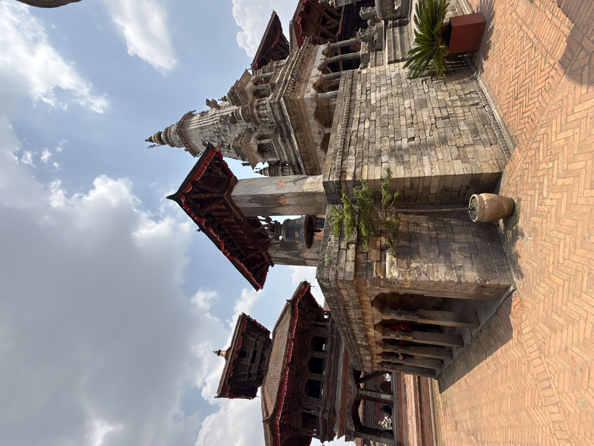 The image showcases a tilted, low-angle view of architectural structures in Bhaktapur, Nepal. Predominantly composed of brick and wood, the buildings feature tiered roofs with red trim. A tall, multi-level spire or pagoda-like structure rises in the background. A large bell hangs from a supporting beam. Vegetation is present on the structures. The foreground depicts a brick-paved area laid in a herringbone pattern. The sky consists of scattered clouds against a blue backdrop.