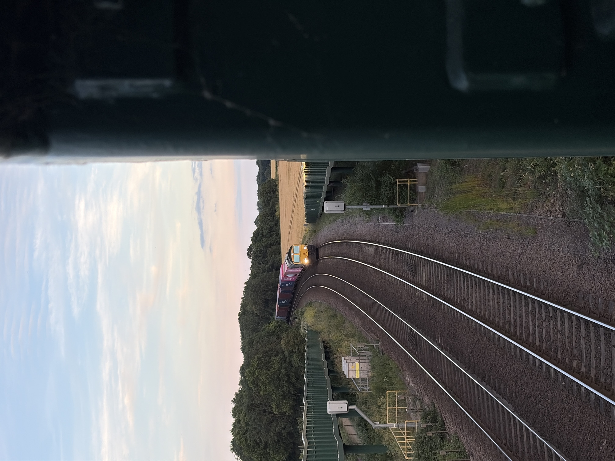 A train with a yellow locomotive and a long line of cargo containers, some colored pink and blue, travels on a curved railway track. The track runs through a rural landscape with a field of crops to the right and a dense line of trees in the background. A green pedestrian bridge with a metal railing extends from the left side of the image, running parallel to the train tracks. The sky is a pale blue with wispy white clouds, tinged with the soft colors of sunset. The overall scene appears to be in East Suffolk, United Kingdom.