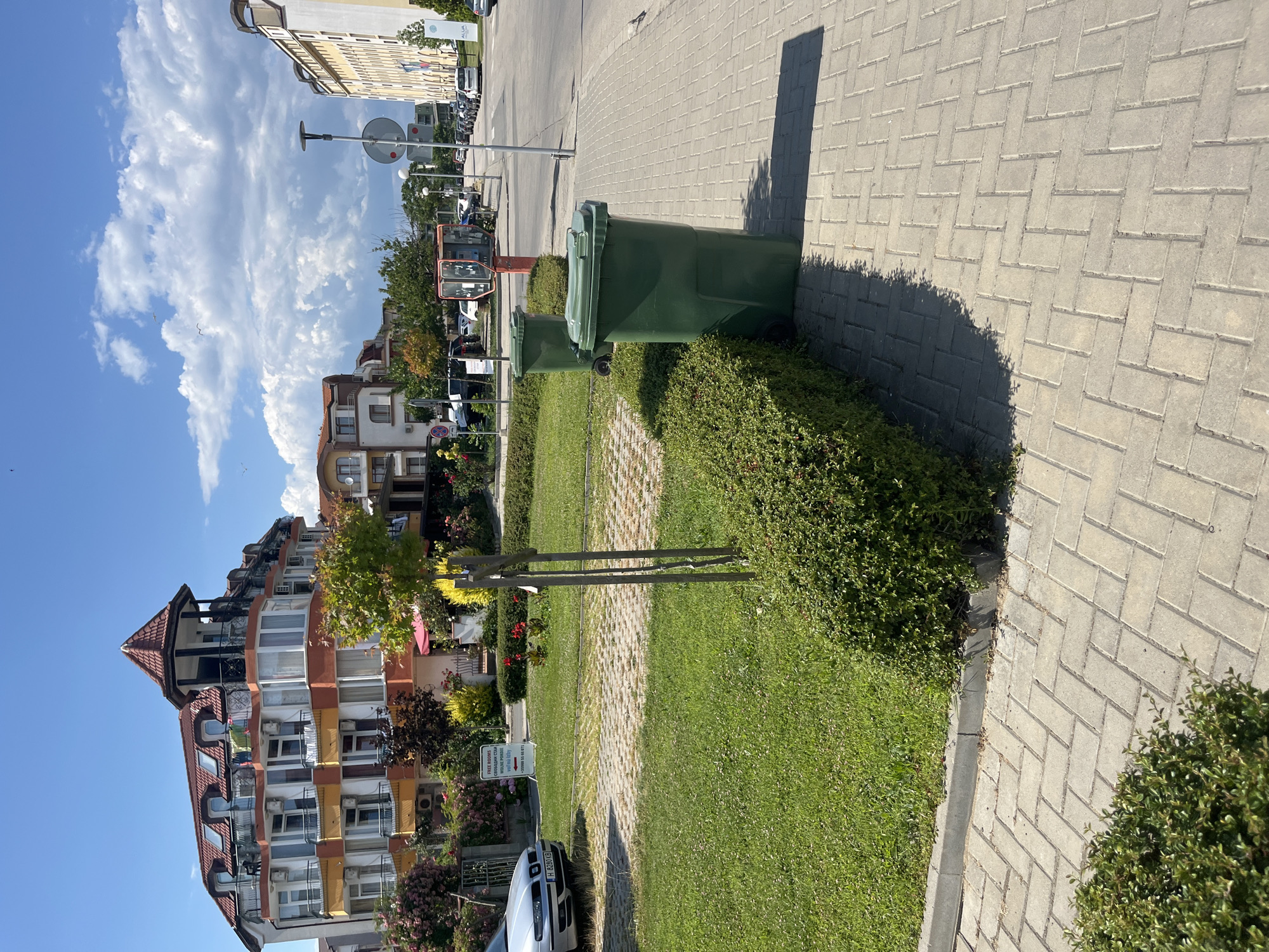 This is a photograph taken in the city of Obzor, Bulgaria, on a clear, sunny day with scattered clouds. The image focuses on a street scene with a residential building in the background and a sidewalk in the foreground.
In the mid-ground, there is a multi-story building with an orange and white facade, featuring balconies and pitched red roofs. Adjacent to this building, there are smaller structures with similar architectural styles. A trimmed green hedge runs along the edge of a grassy area. Two large, dark green plastic trash receptacles are positioned on the paved sidewalk next to the hedge.
To the left of the frame, a silver car with license plate H 8280 KB is parked partially on a textured gray paved surface which appears to be a parking area or walkway. In the distance, a row of parked cars and a light-colored building with a yellow facade are visible along the street. A traffic light and a circular street sign are also present on the right side of the street. A small, weathered wooden post with two crossbars stands in the grassy area.
The sidewalk is paved with light gray rectangular interlocking bricks laid in a herringbone pattern. The grassy area is well-maintained, with short green grass and a few small red flowers. The overall scene depicts a well-kept urban environment, likely a residential or tourist area, given the presence of balconies and the style of the buildings.