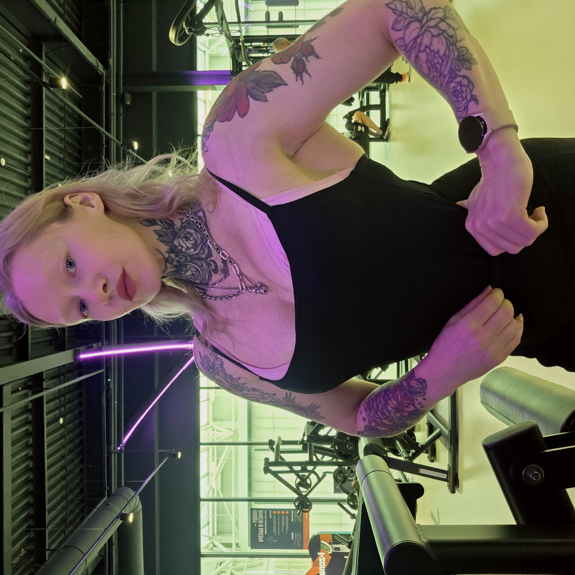 This media file features a person posing in an indoor gym, likely located in Saint Petersburg, Russia. The subject has blonde hair, fair skin, and light-colored eyes, looking directly at the camera with a neutral to slightly contemplative expression. They are wearing a black tank top and a multi-layered silver chain necklace around their neck.
Notable details include extensive tattoos on both arms and their neck, featuring intricate floral and possibly berry designs. A smartwatch with a black face and a light-colored band is visible on their left wrist. The person's right hand is placed on their hip or waist area, and the image is rotated, presenting the subject horizontally.
The background clearly depicts a gym setting with various exercise equipment, including dark metal frames, weights, and padded components. The lighting is artificial and striking, characterized by prominent purple neon lights and a general green ambient light throughout the space. This colorful illumination casts a pinkish glow on the person's skin. The ceiling features an industrial design with corrugated panels, exposed pipes, and additional light fixtures. In the background, a sign with partially visible Cyrillic text, which appears to state "GENERAL RULES," confirms the Russian context. The overall scene suggests a contemporary fitness environment with a distinct neon aesthetic.