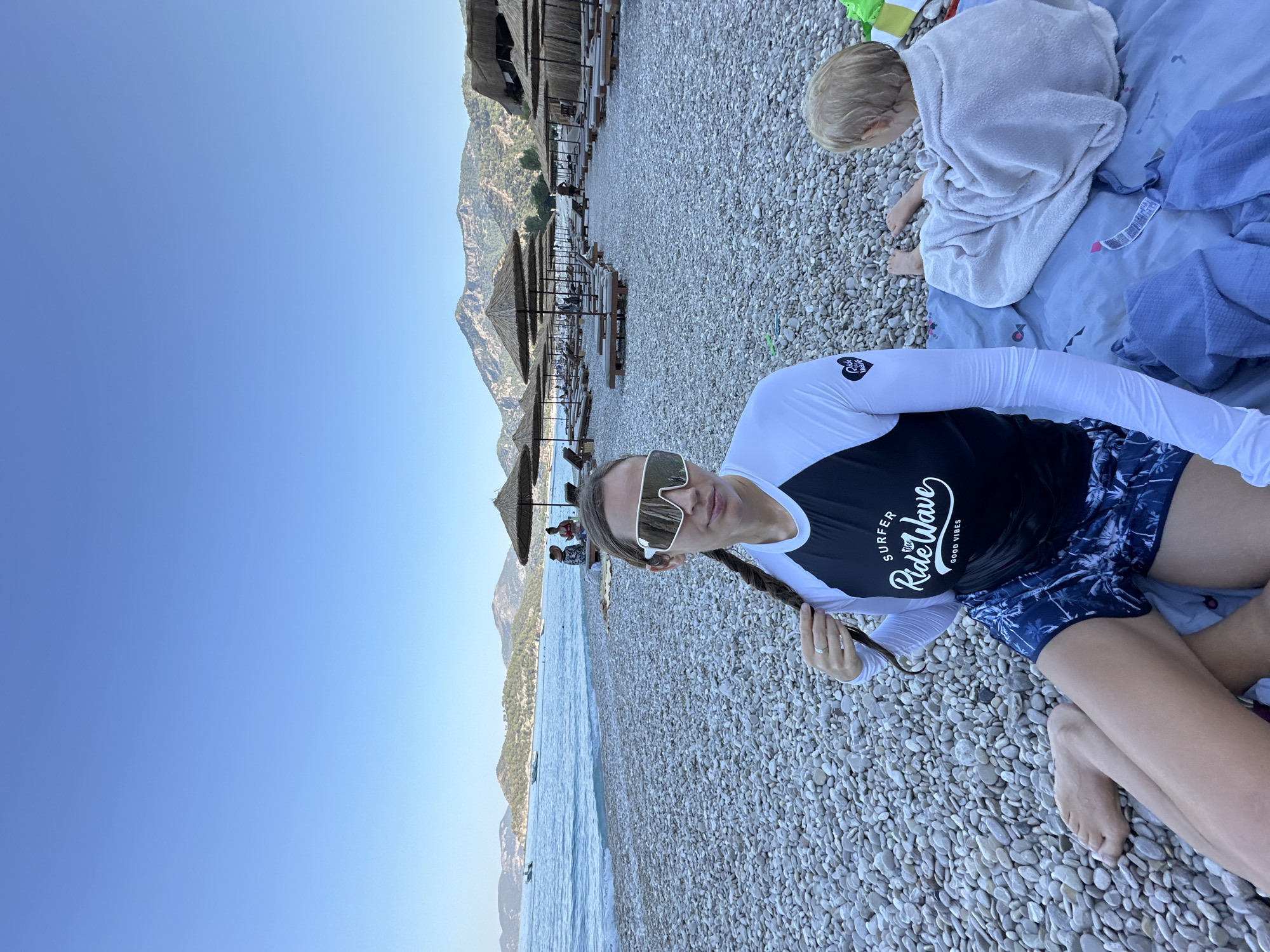 The image depicts a beach scene in Fethiye, Turkey, under a clear blue sky. In the foreground, a woman with a braid, wearing a long-sleeved rash guard with "Surfer Ride Wave Good Vibes" printed on it and patterned swim shorts, sits on a pebble beach, holding her braid. She wears large, white, wraparound sunglasses. To her right, a young child wrapped in a white towel lies on a patterned blanket. The beach is covered in white and gray pebbles, and rows of thatched umbrella-covered sun loungers are set up further back. The sea is visible in the background, with calm water and a few boats. Mountains, covered in greenery, form the backdrop to the scene.