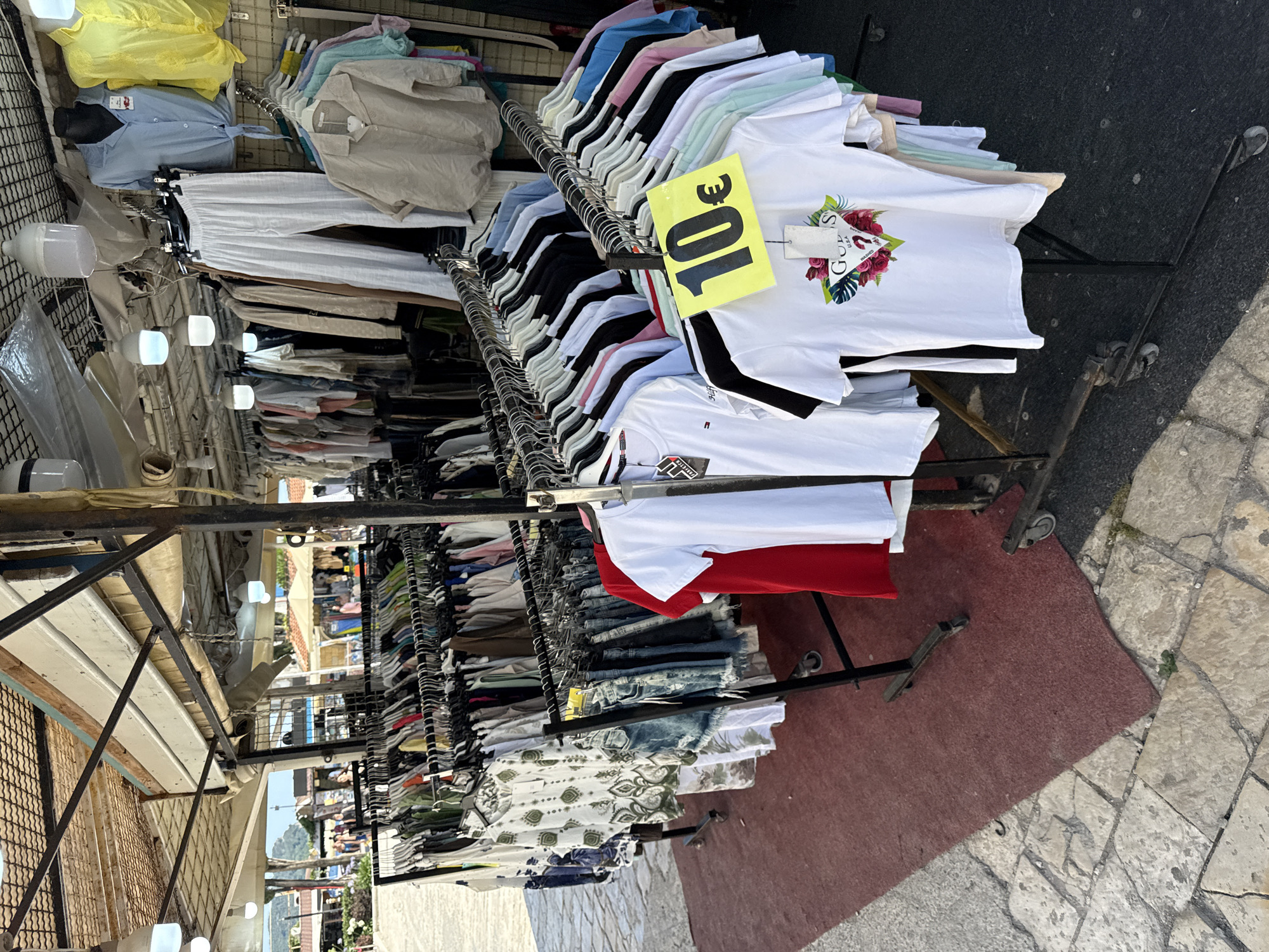 The image depicts an outdoor clothing stall with various garments displayed on racks. The stall appears to be located in a tourist area, possibly in Budva, Montenegro, given the architectural style visible in the background, which includes stone pathways and buildings that are characteristic of coastal Mediterranean towns.
The stall is covered by a canopy, providing shade. In the foreground, racks of clothing are visible, including t-shirts, shorts, and pants. A bright yellow sign with "10€" is prominently displayed on one of the racks. Several t-shirts are neatly hung, some with tags attached. One t-shirt in the foreground features a floral design with the "GUESS" logo.
In the mid-ground, more clothing is arranged on racks, including denim shorts and patterned tops. Further back, the stall extends with more garments hanging. The background reveals a street scene with other stalls, people browsing, and buildings, suggesting a lively market or shopping area. The lighting indicates it is daytime, and the overall atmosphere is casual and commercial.