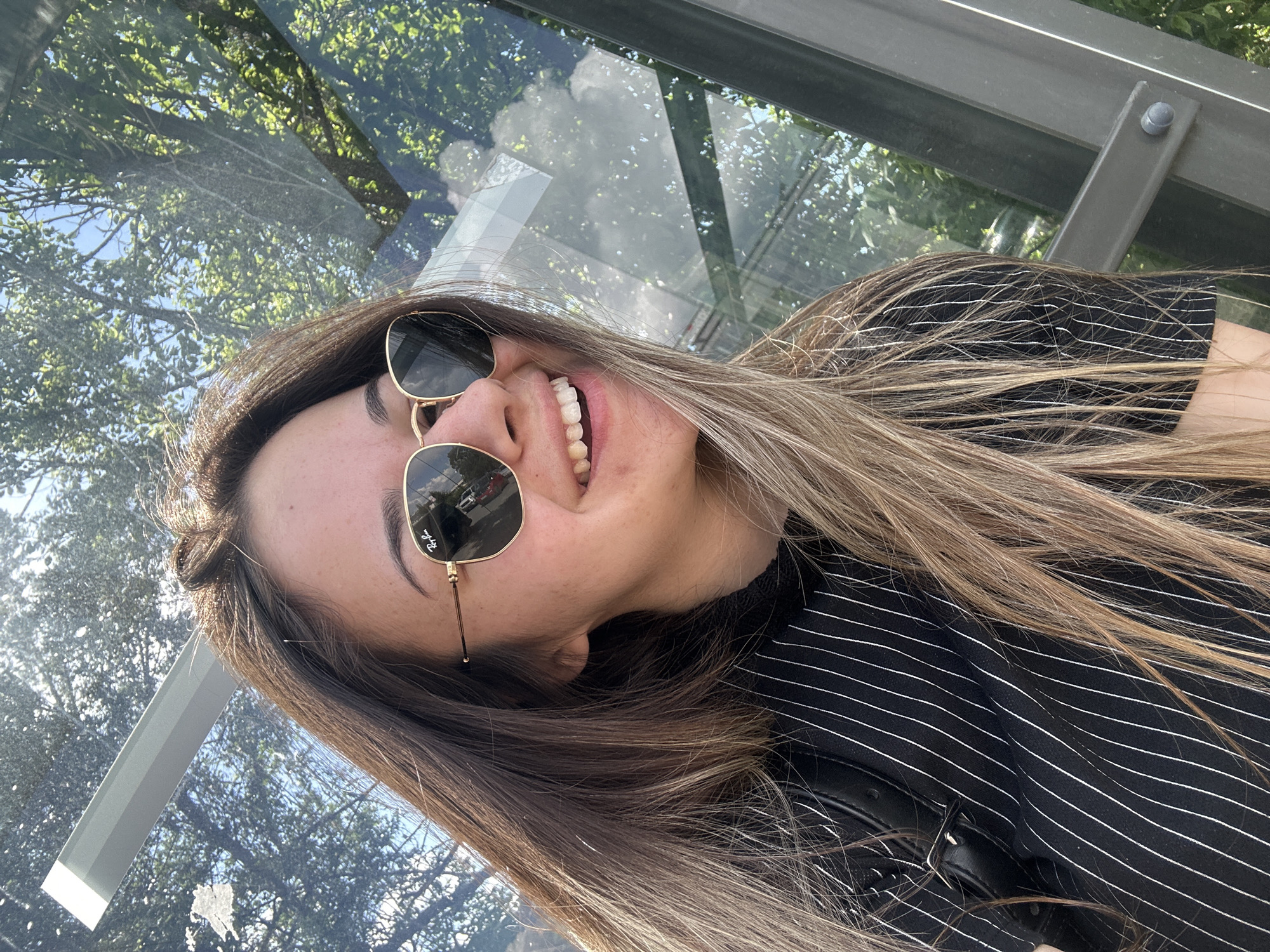 A young woman with long, light brown hair and sunglasses is smiling broadly while standing at a bus stop. The bus stop is made of glass panels held in place by a metal frame. Trees are visible through the glass, reflecting in the surface. The woman is wearing a black and white striped shirt and possibly a jacket. The location is difficult to determine without more context, but the bus stop design and overall environment suggest it could be Sofia, Bulgaria.