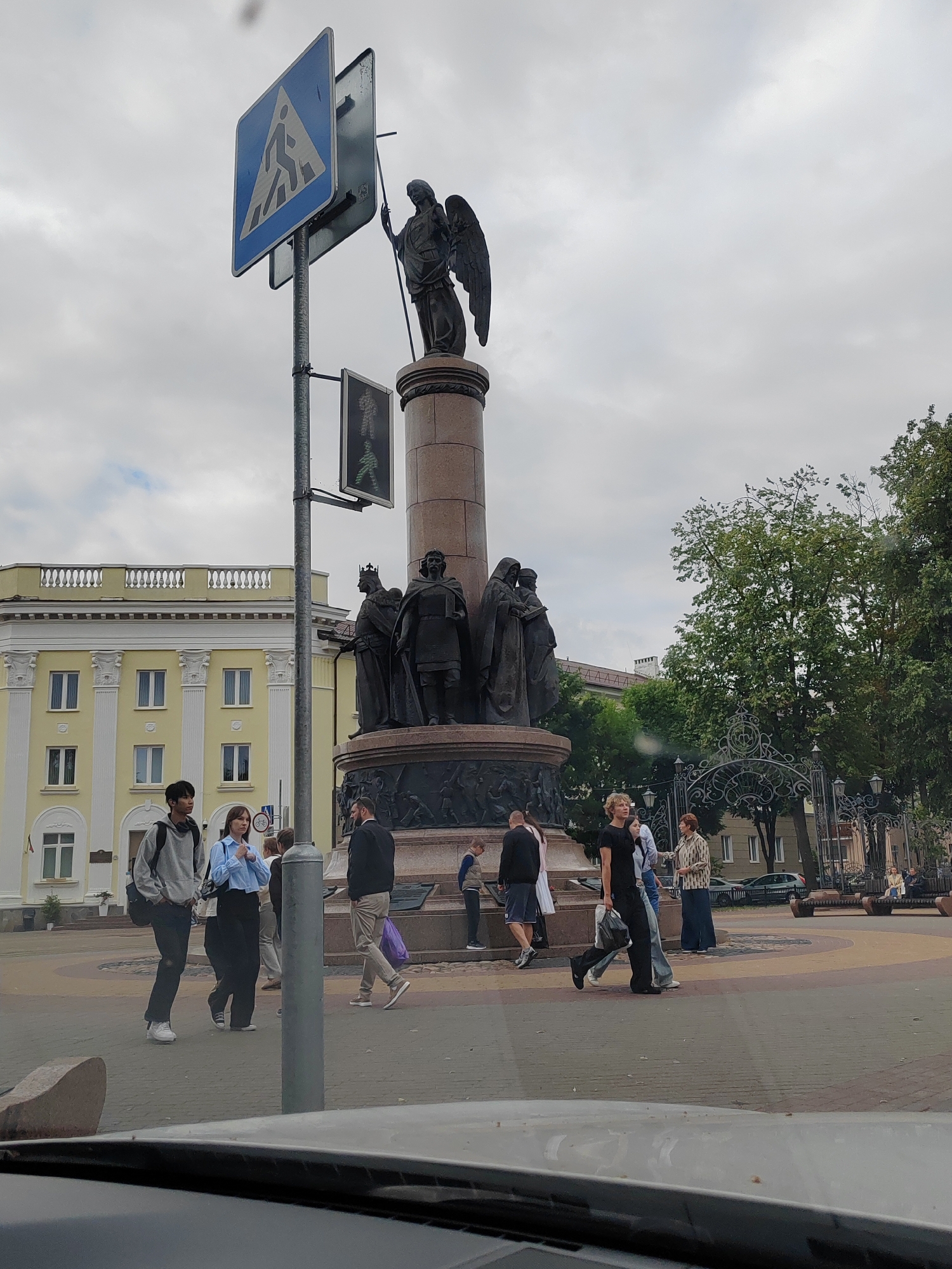 The image depicts a scene in Brest, Belarus, focusing on a prominent monument situated in an open urban space. The monument itself is a tall, cylindrical structure adorned with several bronze statues of historical figures, topped by a winged angel holding a staff. Adjacent to the monument is a pedestrian crossing sign and a traffic light displaying a green walking figure.
Several individuals are visible in the vicinity. Two young people, a male with a backpack and a female in a blue shirt, are walking on the paved area in the foreground. Other pedestrians are scattered throughout the scene, some walking, others standing near the monument or benches. In the background, a light yellow building with white decorative elements and numerous windows is visible, suggesting it might be a public building or administrative office. Green trees and ornate metal fencing add to the urban landscape. The sky is overcast, indicating a cloudy day. The photograph appears to have been taken from a slightly elevated perspective, possibly from a vehicle, as the lower edge of the image shows the dashboard or windshield of a car.