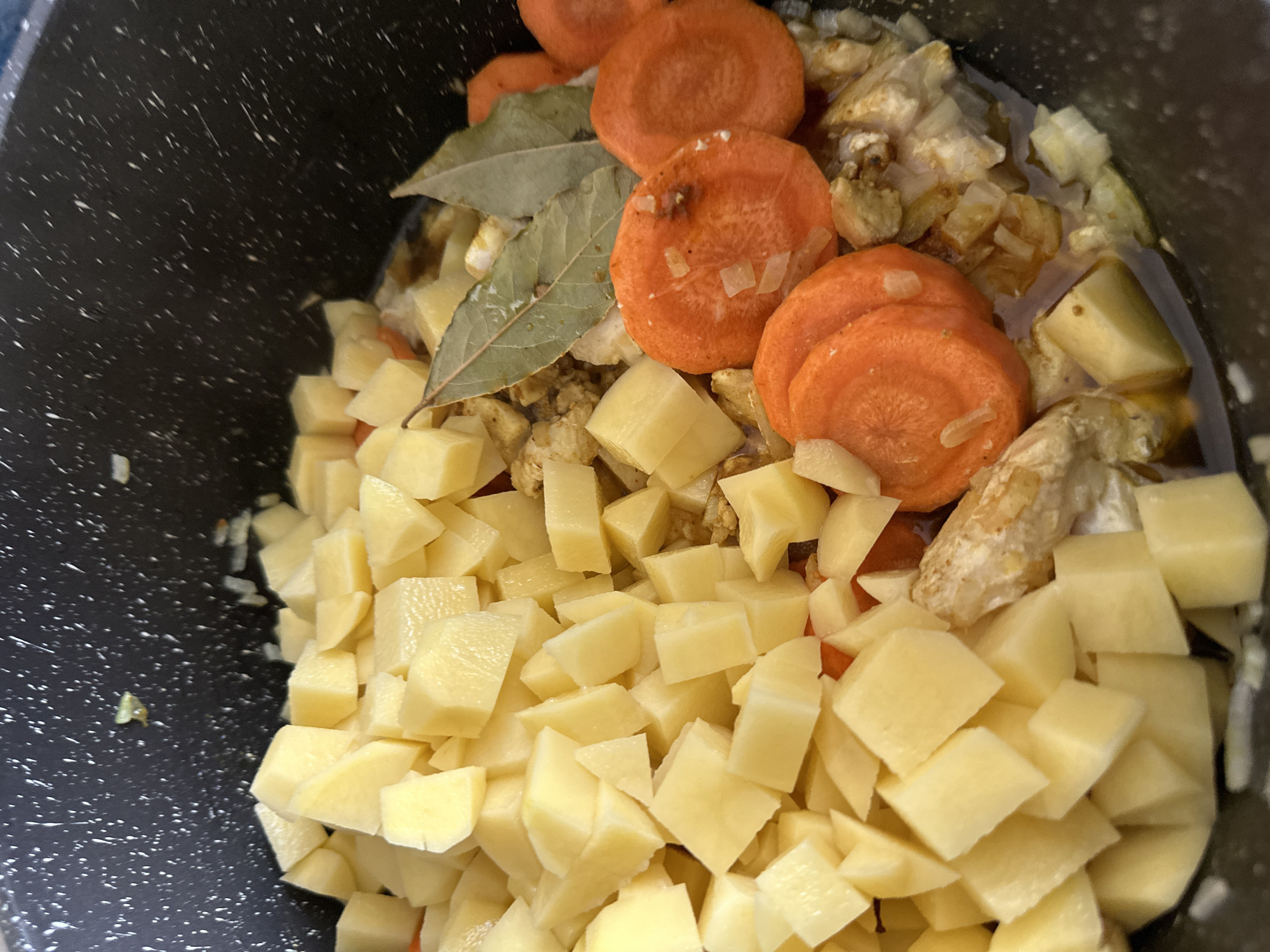 The image shows a close-up view of a black pot filled with ingredients for a stew or soup. Visible are diced potatoes, sliced carrots, onions, and bay leaves. The food is inside an indistinguishable environment.