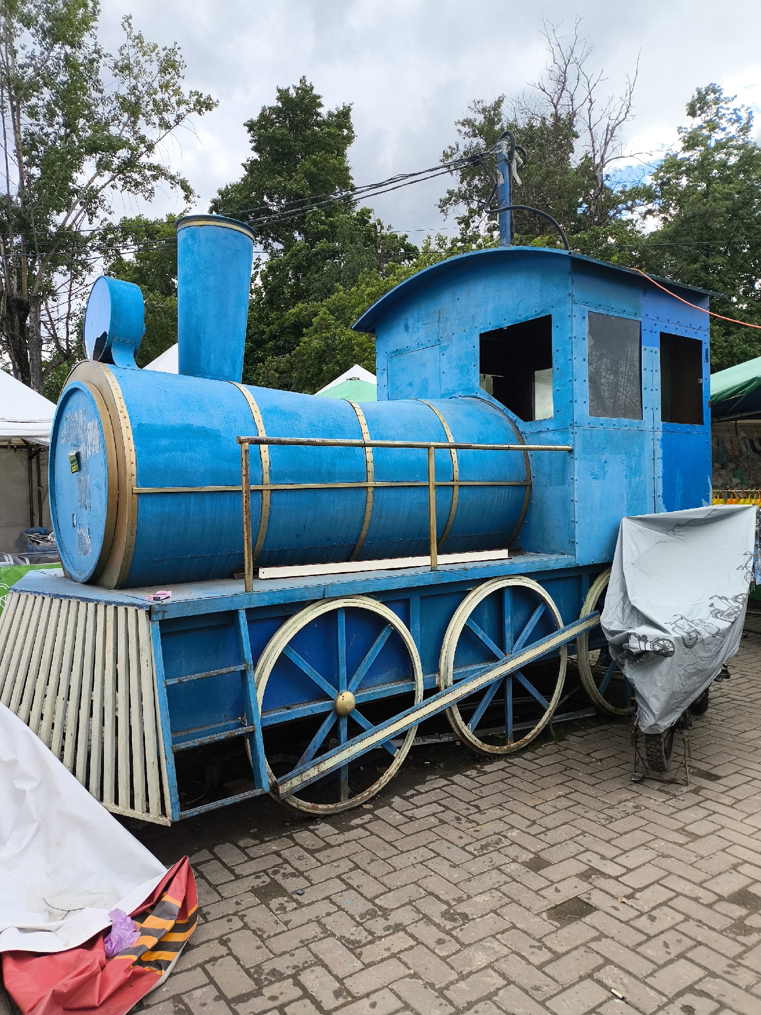 A blue, non-functional locomotive is stationary on a brick-paved surface. The locomotive features large, painted wheels and a front grill made of vertical white and light-yellow bars. It has a cylindrical body with gold-colored trim. The cab of the train has empty window openings. A light-gray cover drapes over an object situated immediately to the right of the train. In the background, trees are visible along with parts of tents or canopies. The sky is overcast. The location is Unknown, Unknown.